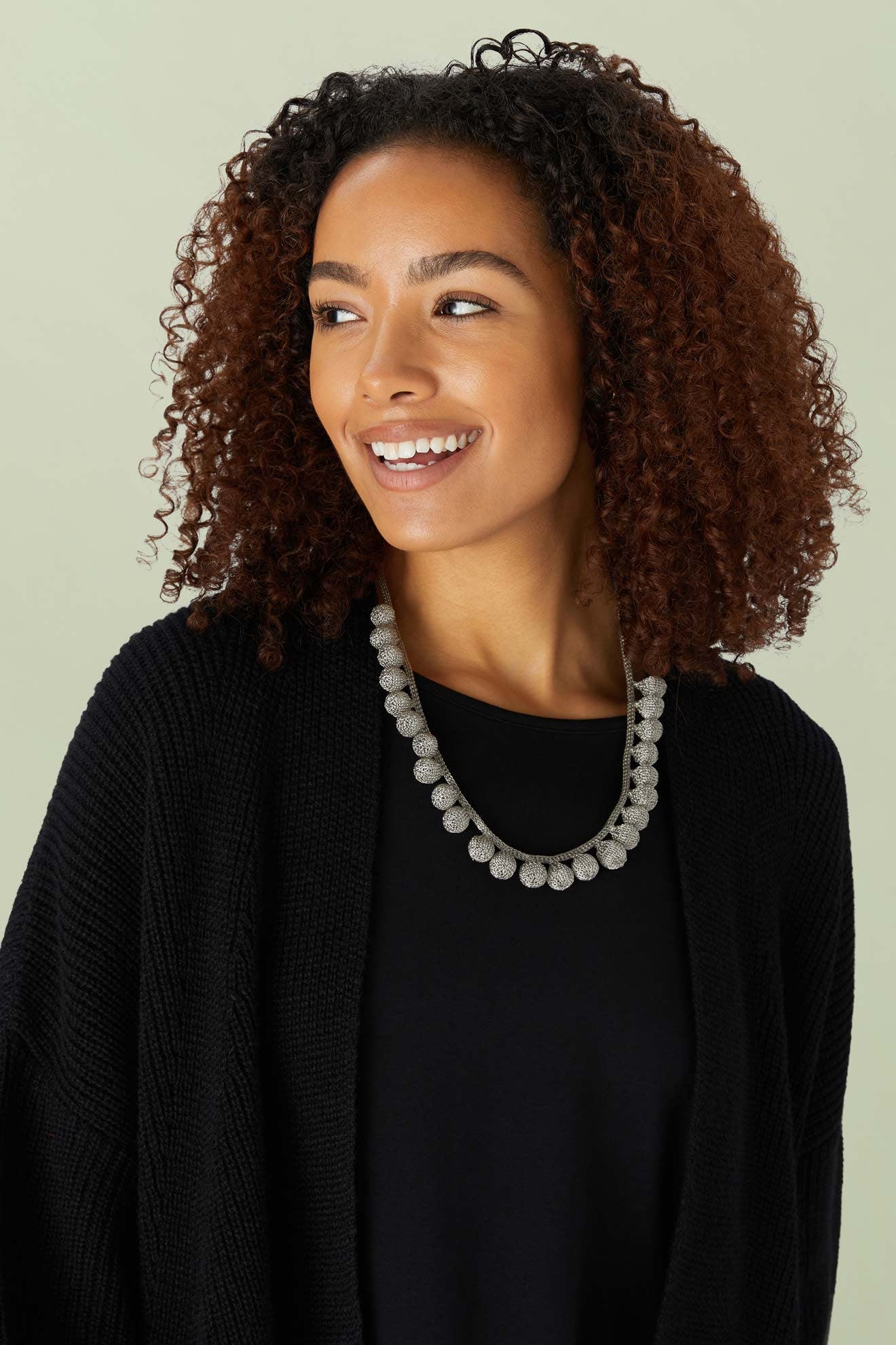 Woman with curly hair wearing a black top and cardigan, smiling and accessorized with the Crochet Tie Necklace, a chunky handcrafted piece inspired by Balinese jewellery, against a plain background.