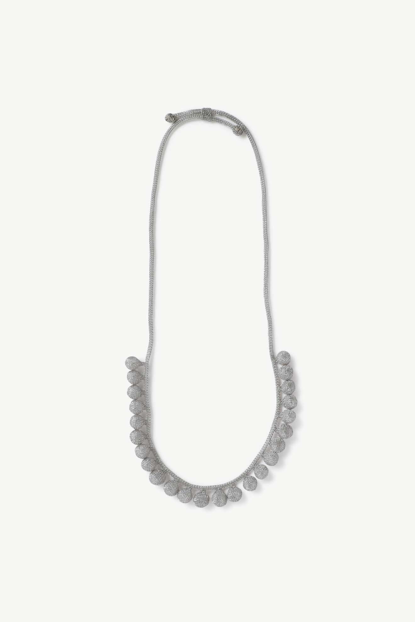 The Crochet Tie Necklace is a handcrafted gray cord piece with round, textured beads spaced evenly along the lower half, inspired by Balinese jewelry.