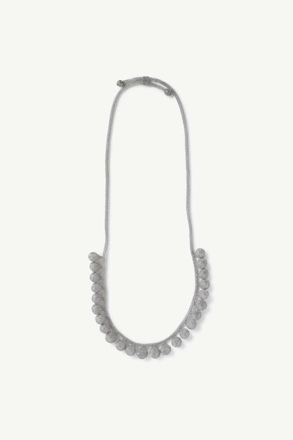 The Crochet Tie Necklace is a handcrafted gray cord piece with round, textured beads spaced evenly along the lower half, inspired by Balinese jewelry.