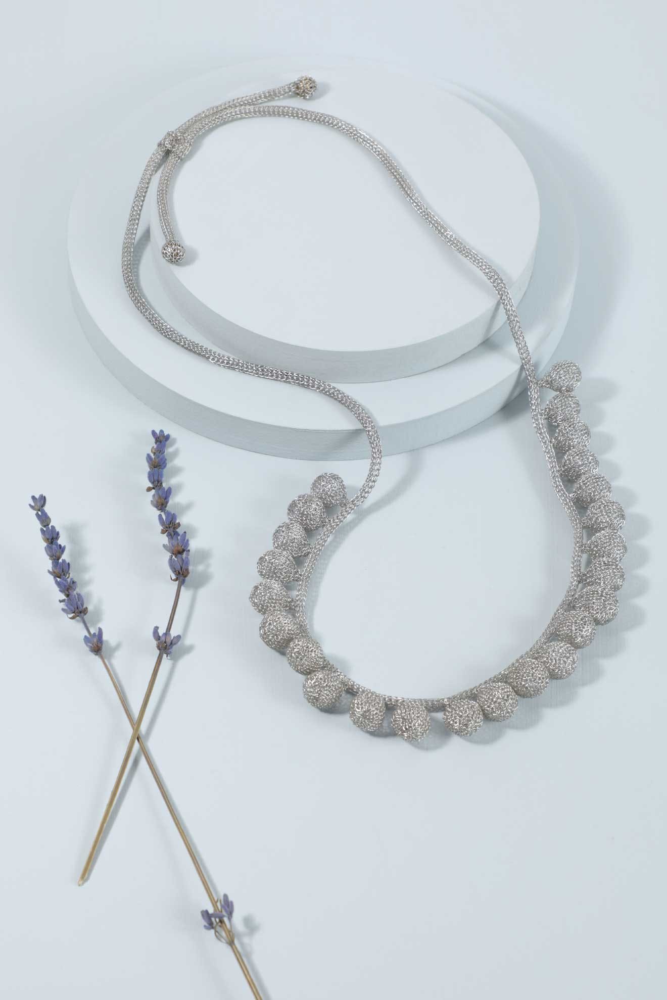The Crochet Tie Necklace, featuring silver handcrafted leaf-shaped charms and delicate wire wrapping, is displayed on a white circular platform beside two sprigs of dried lavender.