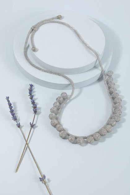 The Crochet Tie Necklace, featuring silver handcrafted leaf-shaped charms and delicate wire wrapping, is displayed on a white circular platform beside two sprigs of dried lavender.