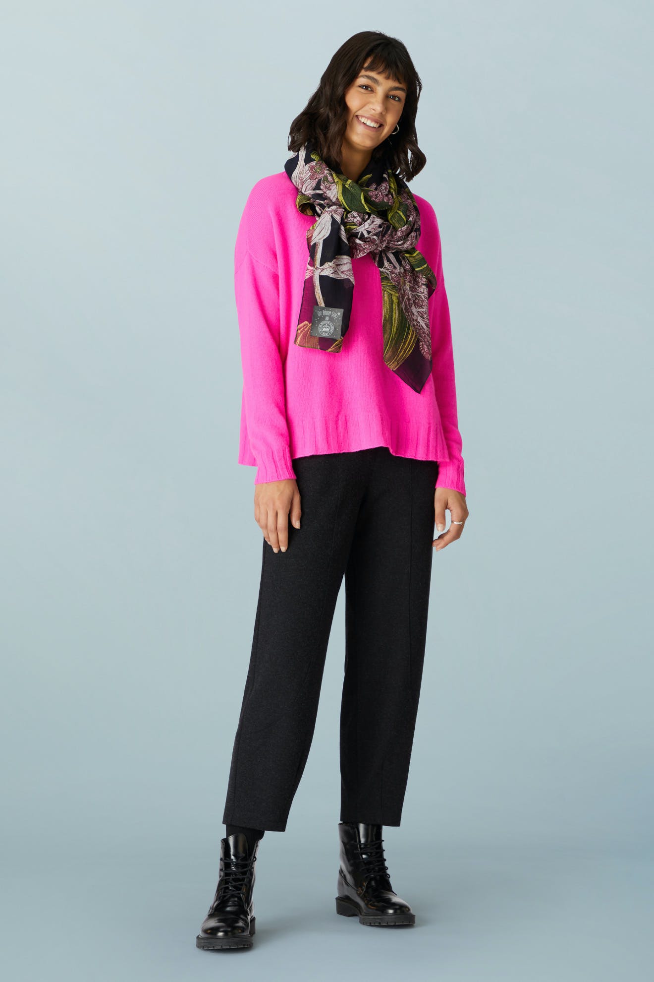 A woman wears a bright pink sweater, a patterned scarf, and black AW21 Marl Jersey Bubble Trouser with black lace-up boots.