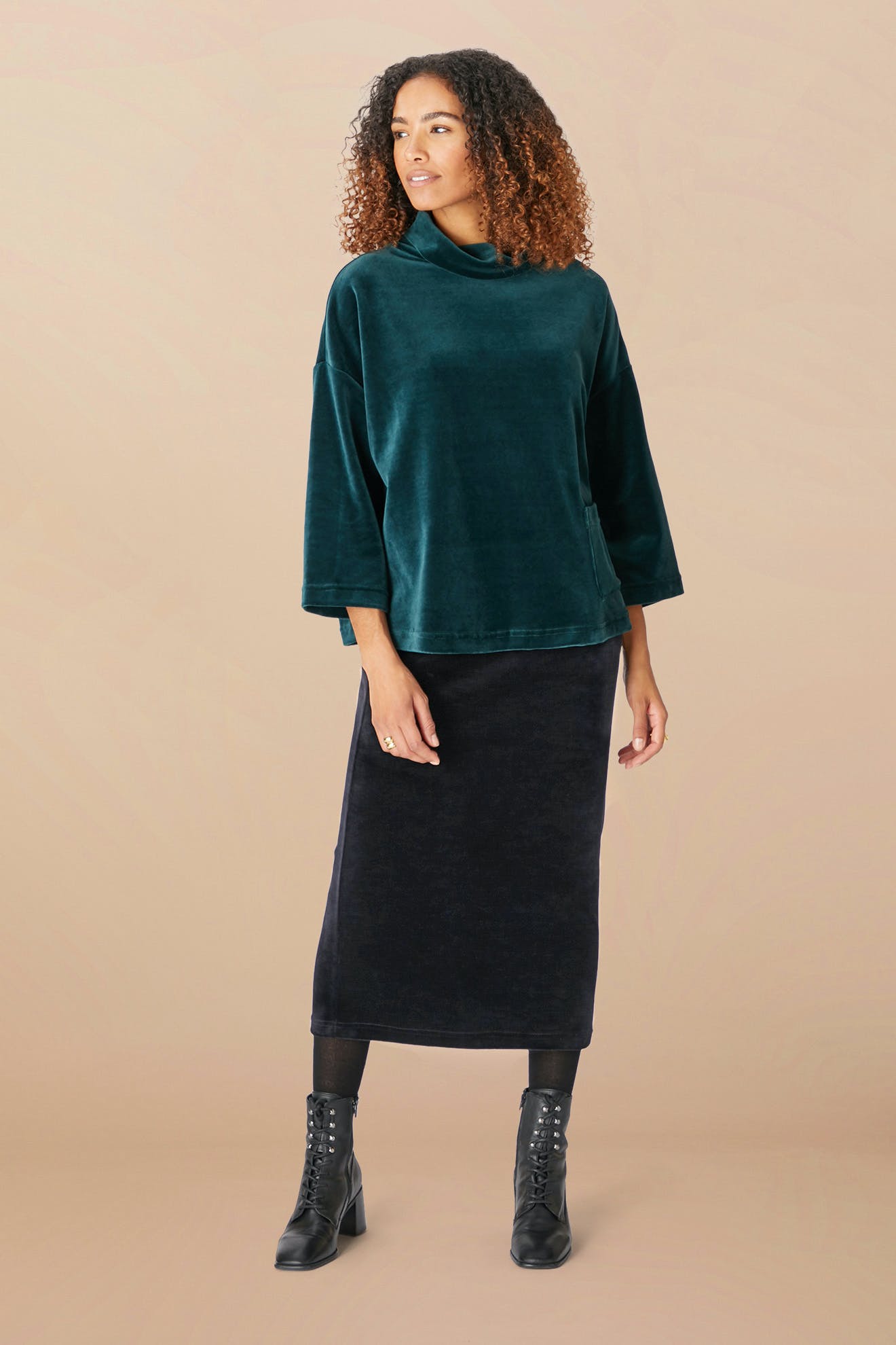 A woman stands against a beige background wearing a dark green velvet top, the Velvet Jersey Tube Skirt in black, high-quality black tights, and black ankle boots. She has curly hair and is looking slightly to her right.