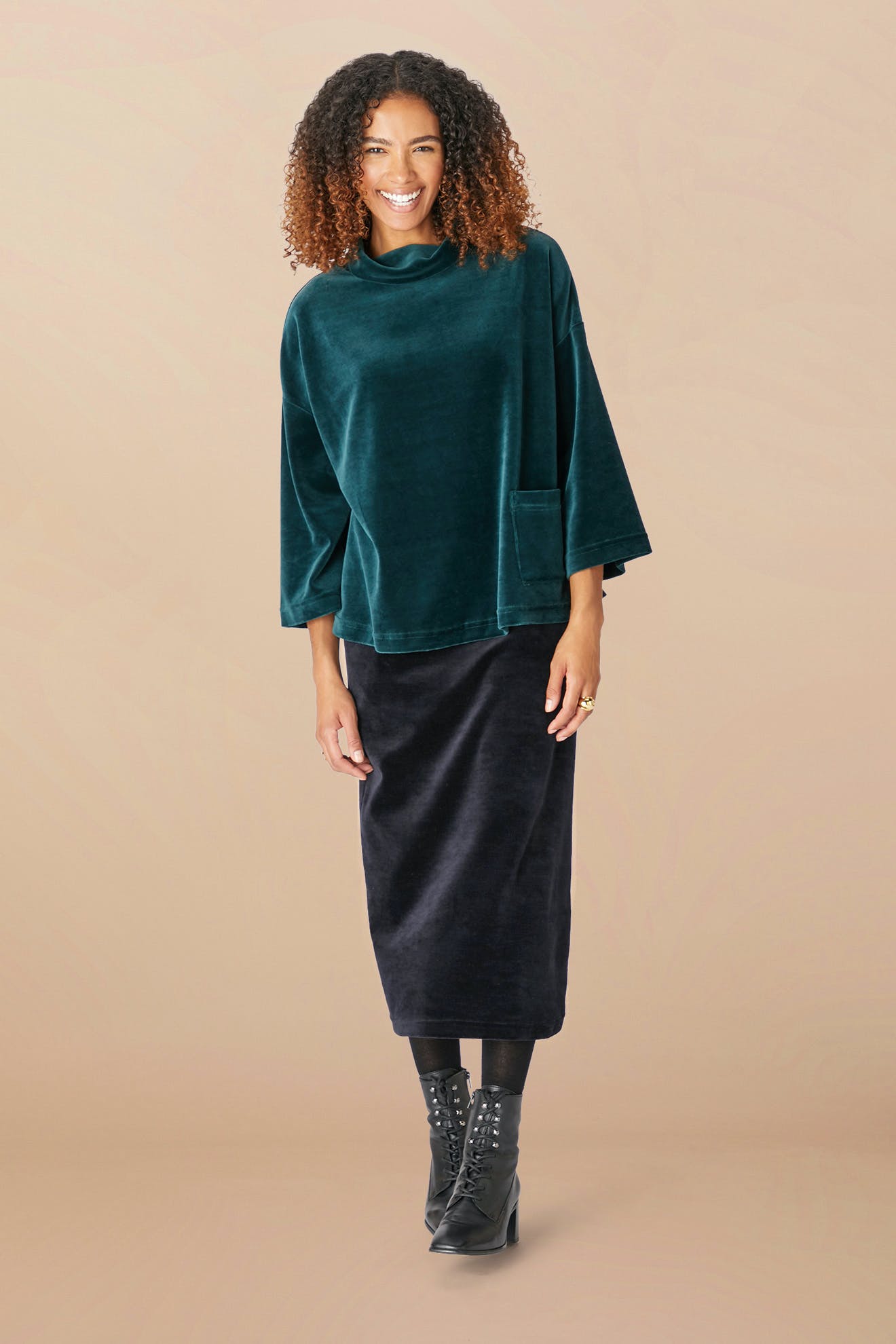 A person with curly hair is smiling and wearing a loose, dark green top and a long black Velvet Jersey Tube Skirt with a classic silhouette. They are also wearing black ankle boots and standing against a plain beige background.