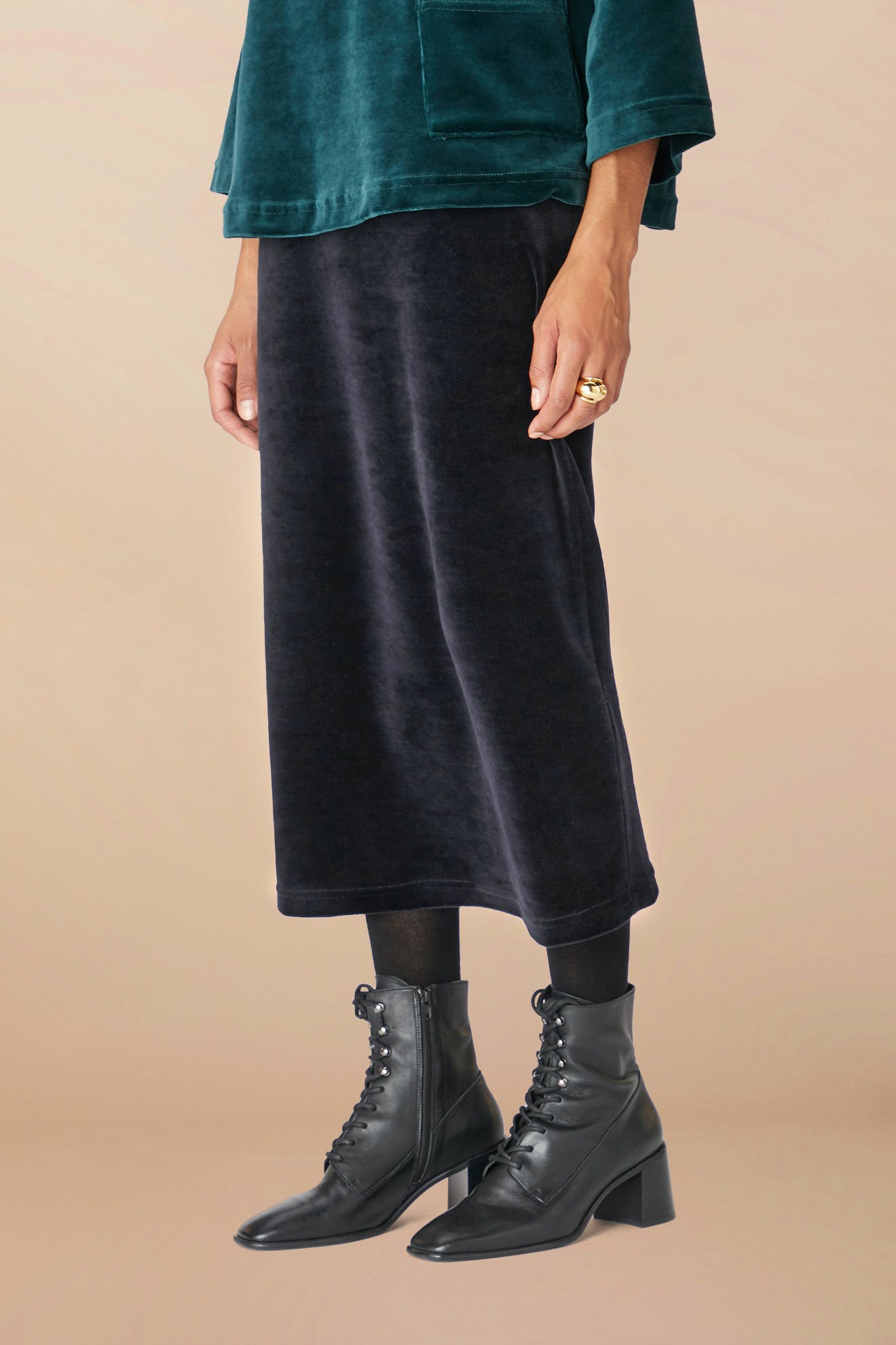 Person wearing a green top, Velvet Jersey Tube Skirt in black velvet, black tights, and black lace-up ankle boots stands with one hand visible.