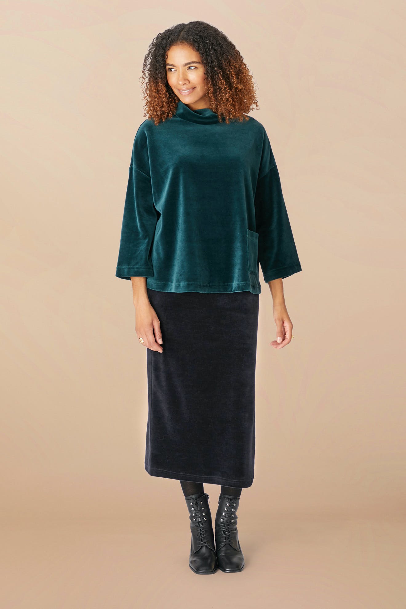 A person with curly hair wearing a dark green long-sleeve top and a black Velvet Jersey Tube Skirt stands against a beige background, embodying a classic silhouette.