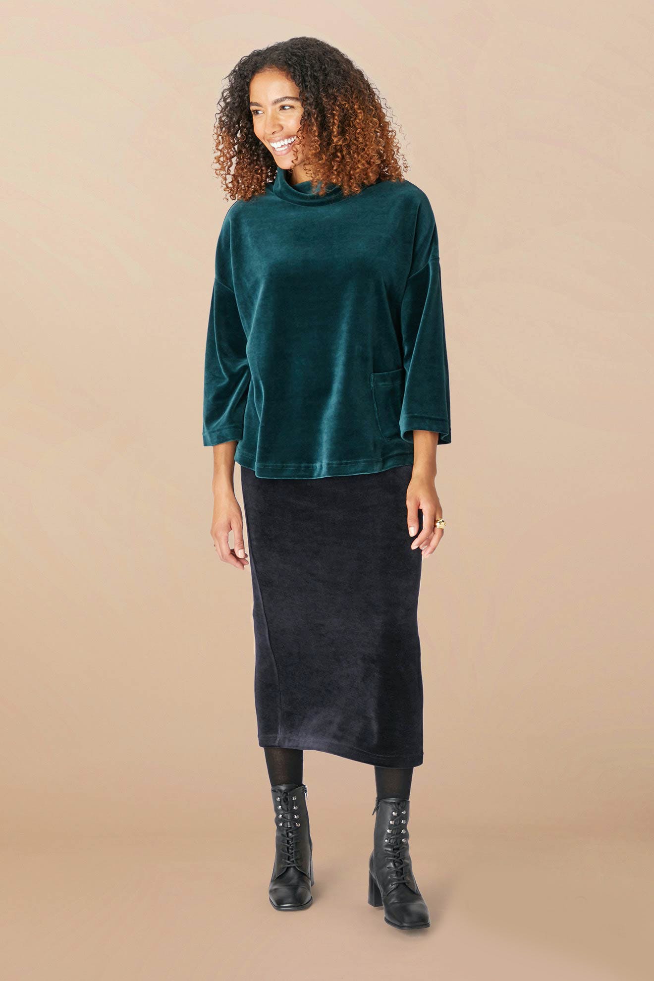 A woman with curly hair, dressed in a dark green long-sleeved top and a classic black Velvet Jersey Tube Skirt, stands smiling against a plain beige background.