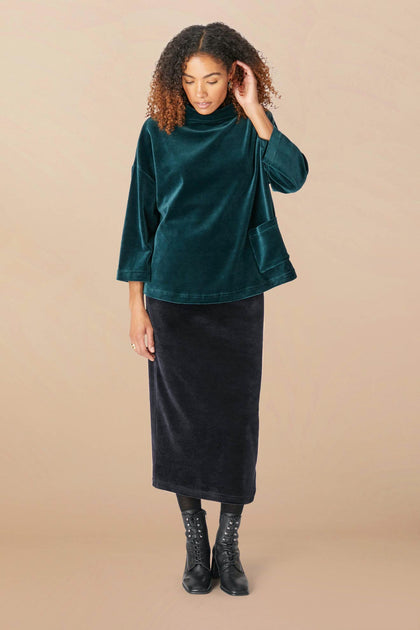 A person with curly hair is wearing a dark green velvet top, a Velvet Jersey Tube Skirt that enhances the classic silhouette, and black lace-up boots, standing against a plain beige background.