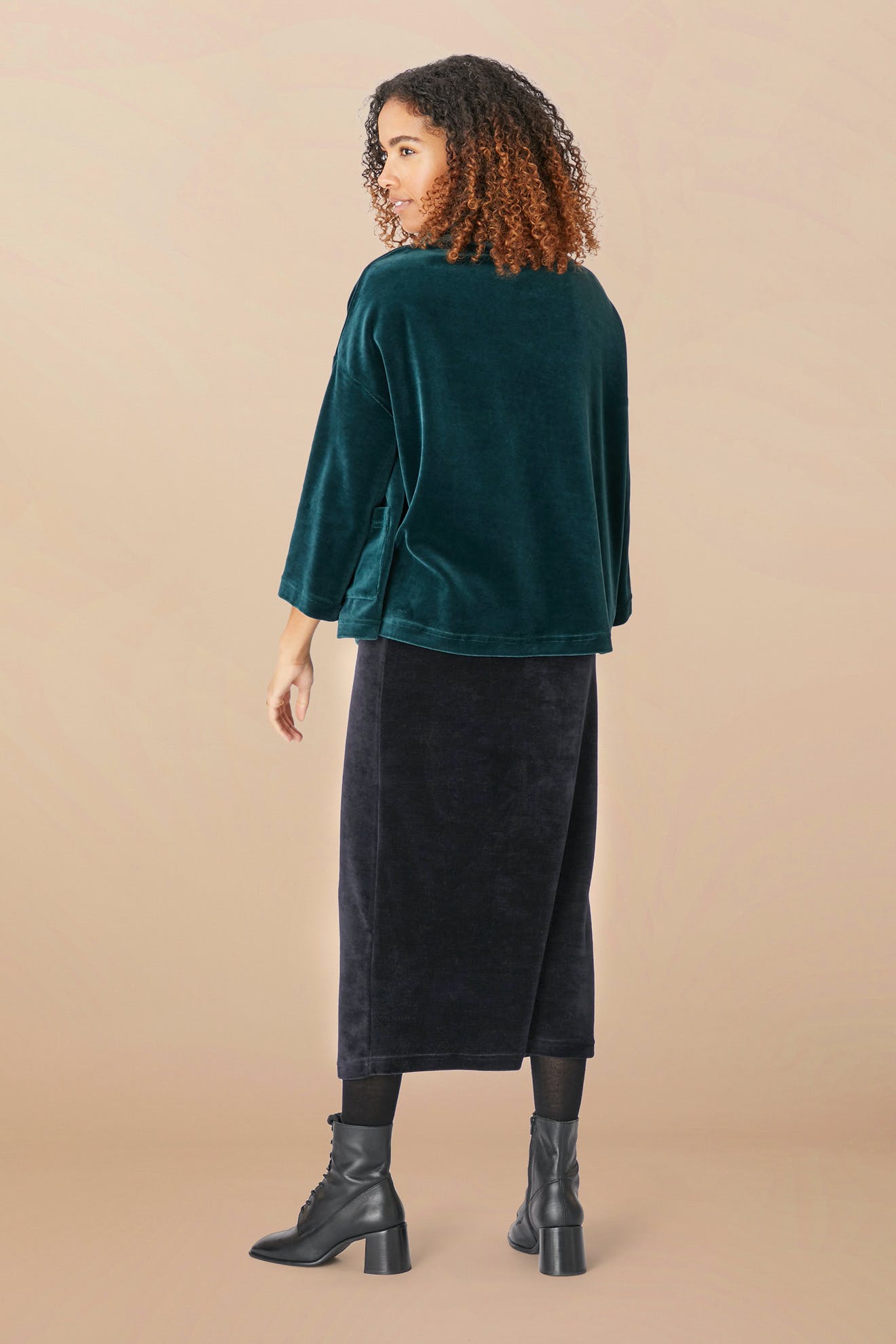 A person with curly hair stands sideways, wearing a dark green, loose-fitting top, a classic silhouette Velvet Jersey Tube Skirt, and black boots against a beige background.