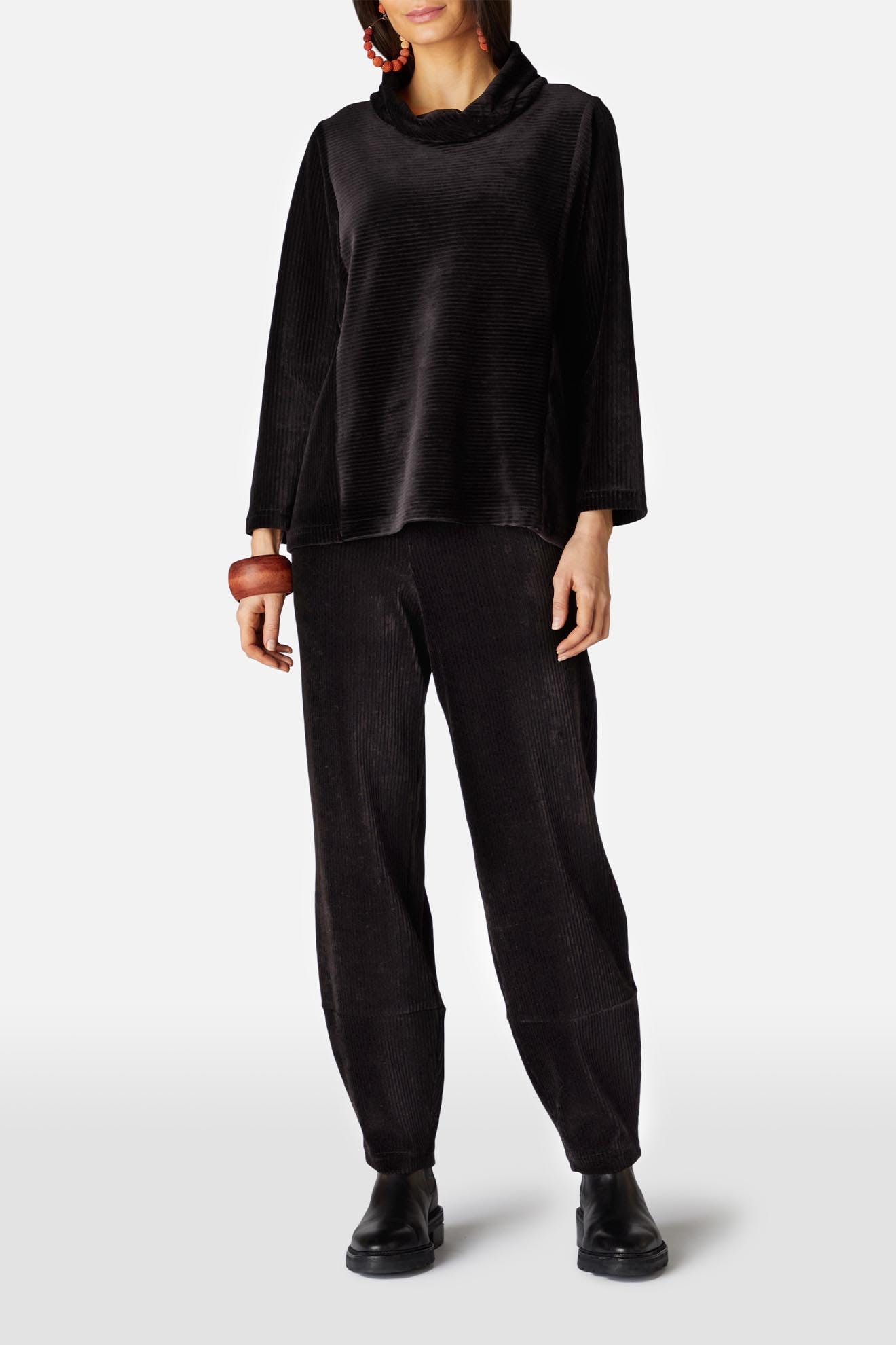 A person wearing the Velvet Rib Jersey Top, velvet jersey slim trousers, black shoes, a red earring, and a wooden bracelet is standing against a plain white background.