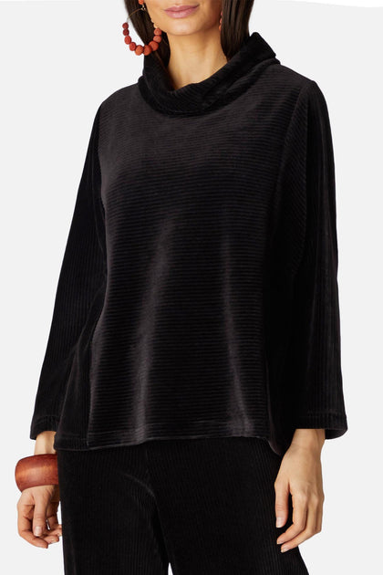 A person wearing the Velvet Rib Jersey Top in black, with long sleeves and a cowl neck, paired with matching Velvet Jersey Slim Trousers, and holding a wooden clutch.