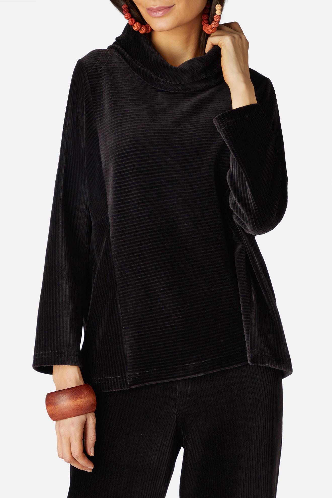 A person is wearing a black corduroy outfit with a Velvet Rib Jersey Top and Velvet Jersey Slim Trousers. They have dangling beaded earrings and a wooden bracelet. The background is plain white.