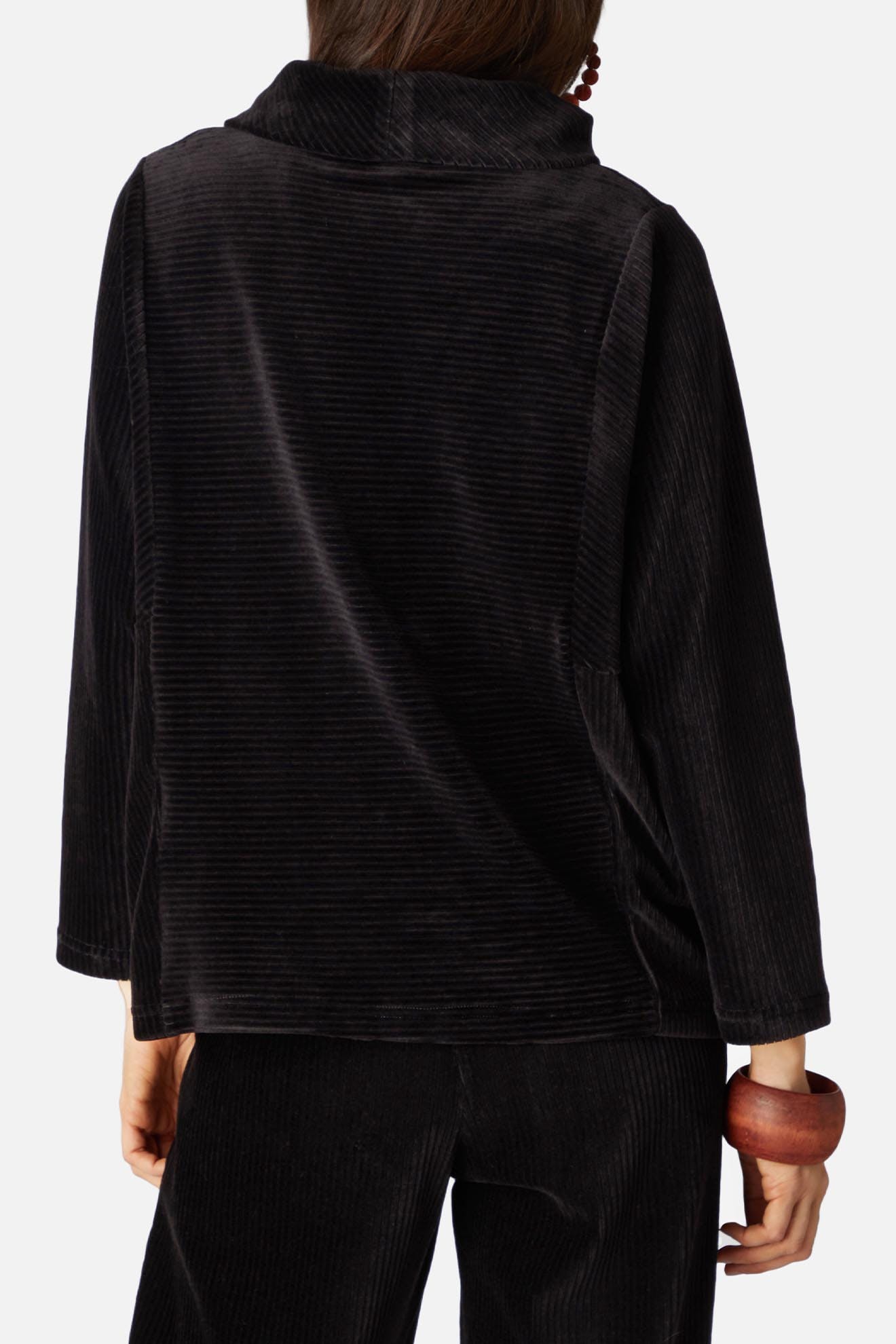 A person is shown from the back wearing a black, long-sleeved, velvet rib jersey top with a cowl neck. They are also wearing matching trousers and holding a wooden bangle in their left hand.