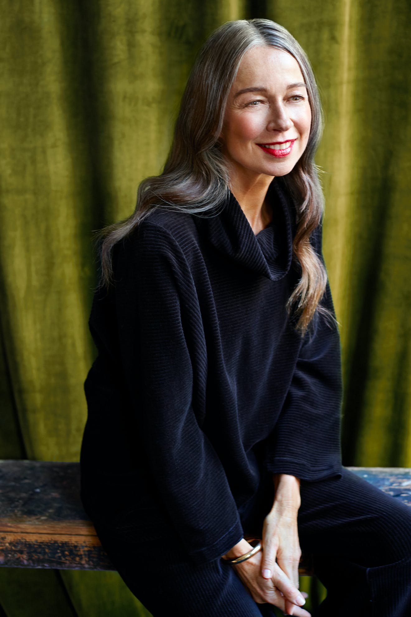 Smiling woman with long, gray-streaked hair, wearing a black cowl neck sweater and Velvet Rib Jersey Top, sits on a bench in front of a green curtain.