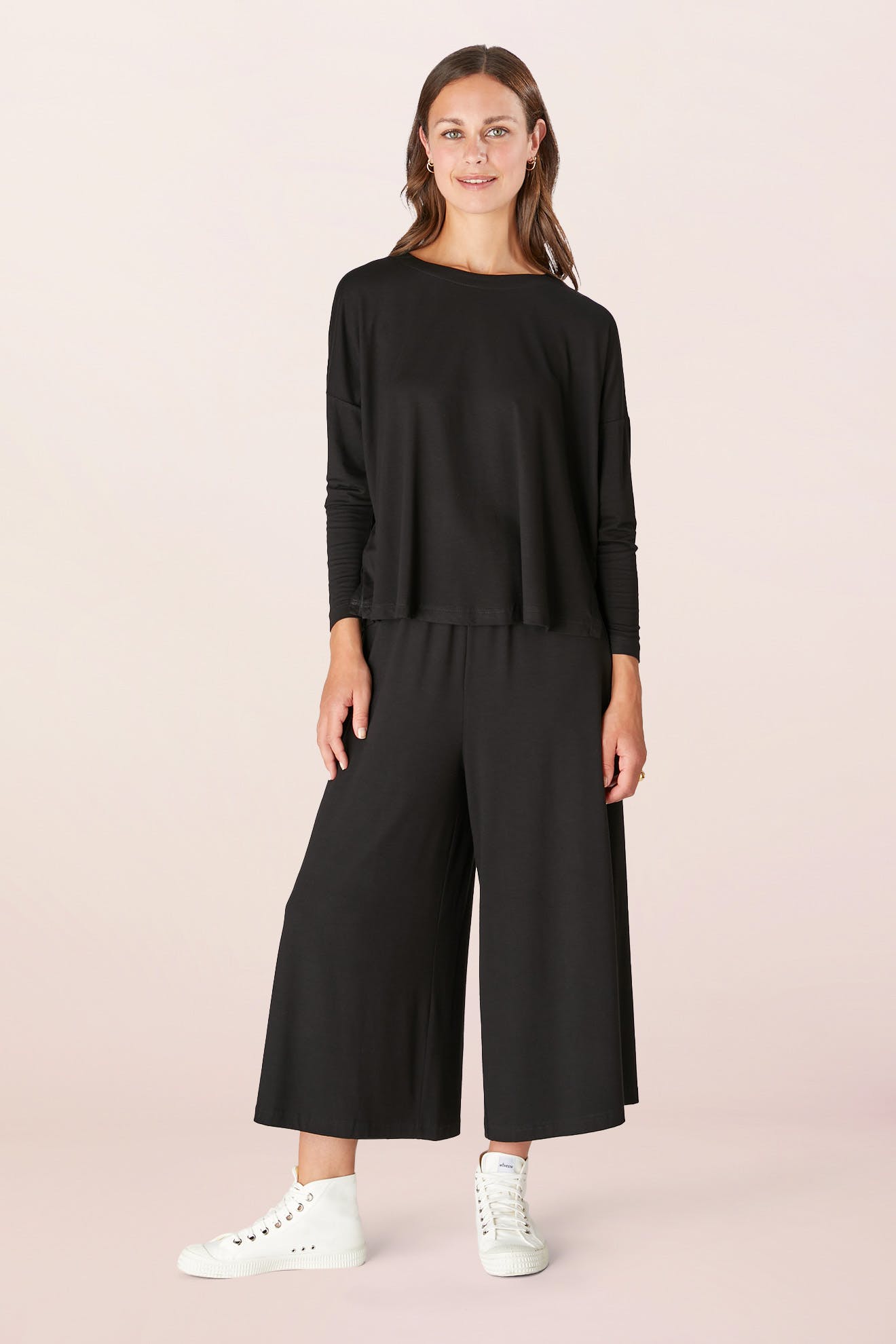 A person wearing a Fluid Crepe Jersey Relaxed Top paired with wide-leg pants and white sneakers, standing and smiling against a plain background.