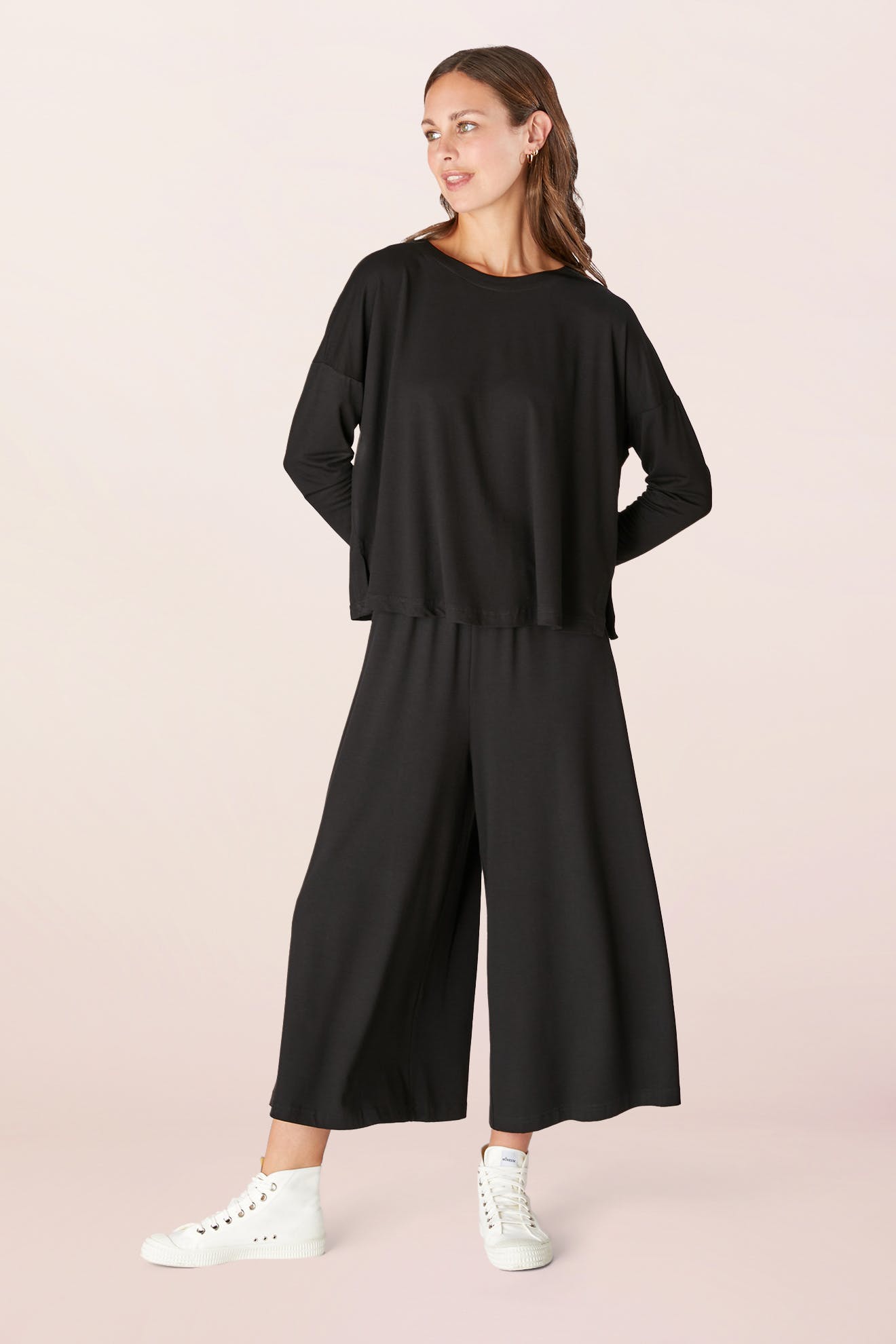 A person wearing the Fluid Crepe Jersey Relaxed Top in black and wide-leg pants with an A-line silhouette poses against a neutral background. They complete the look with white sneakers, exuding effortless style.