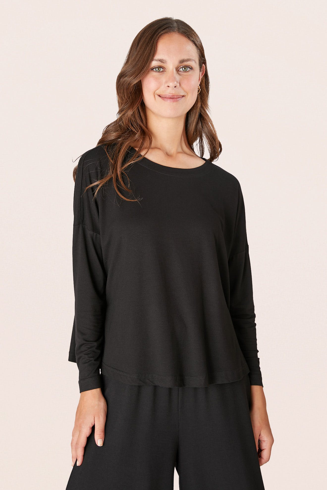 A person stands smiling, wearing a Fluid Crepe Jersey Relaxed Top in black, paired with fluid crepe pants against a plain background.