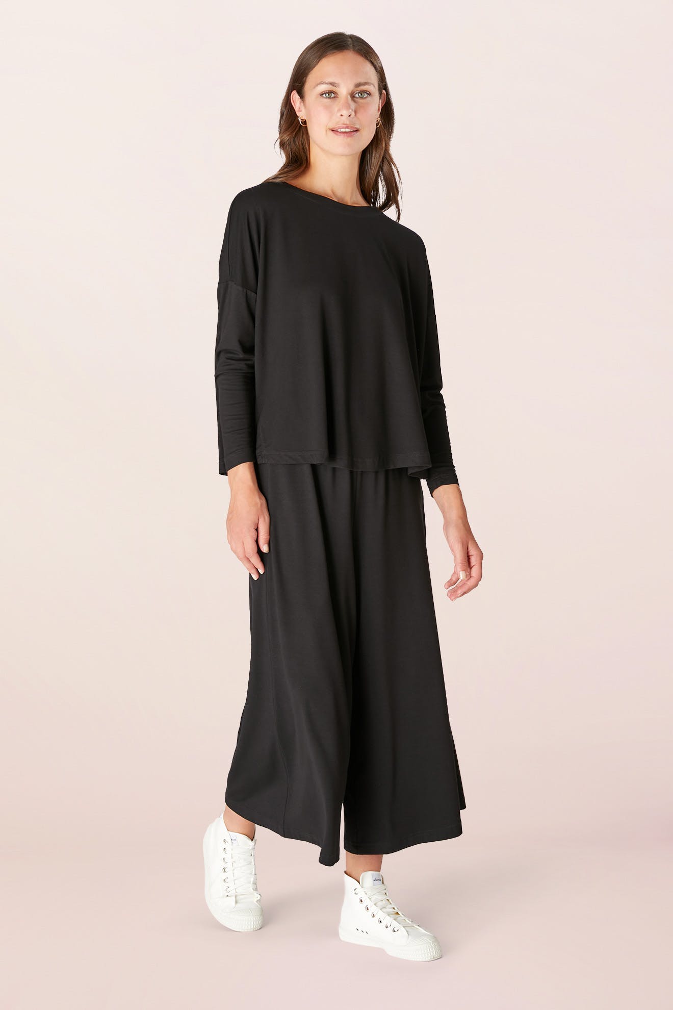 A person dressed in a Fluid Crepe Jersey Relaxed Top and white sneakers stands against a plain background, effortlessly showcasing an A-line silhouette.
