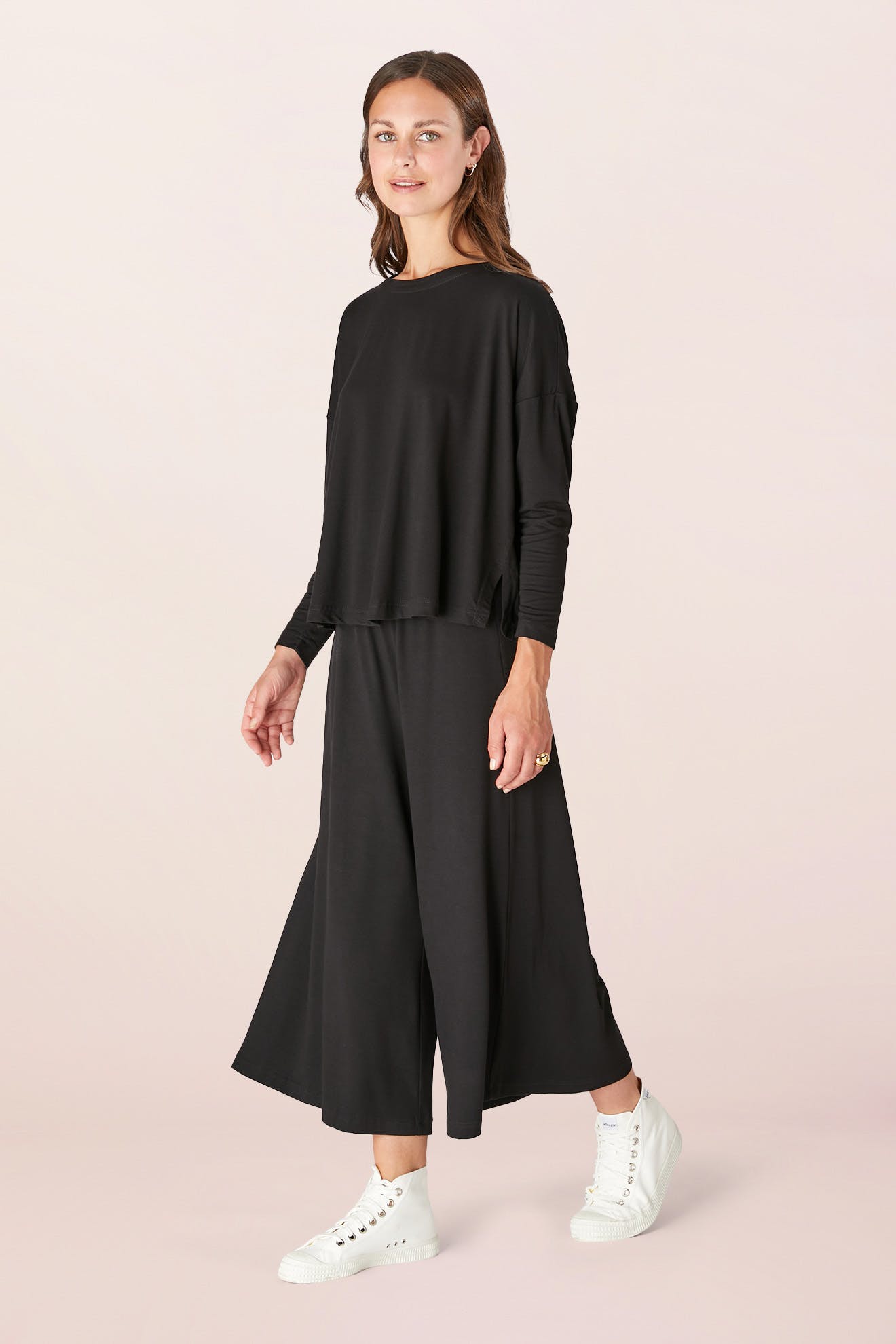 Wearing a Fluid Crepe Jersey Relaxed Top in black and wide-leg pants, a woman glides gracefully, her white sneakers adding a touch of casual elegance.