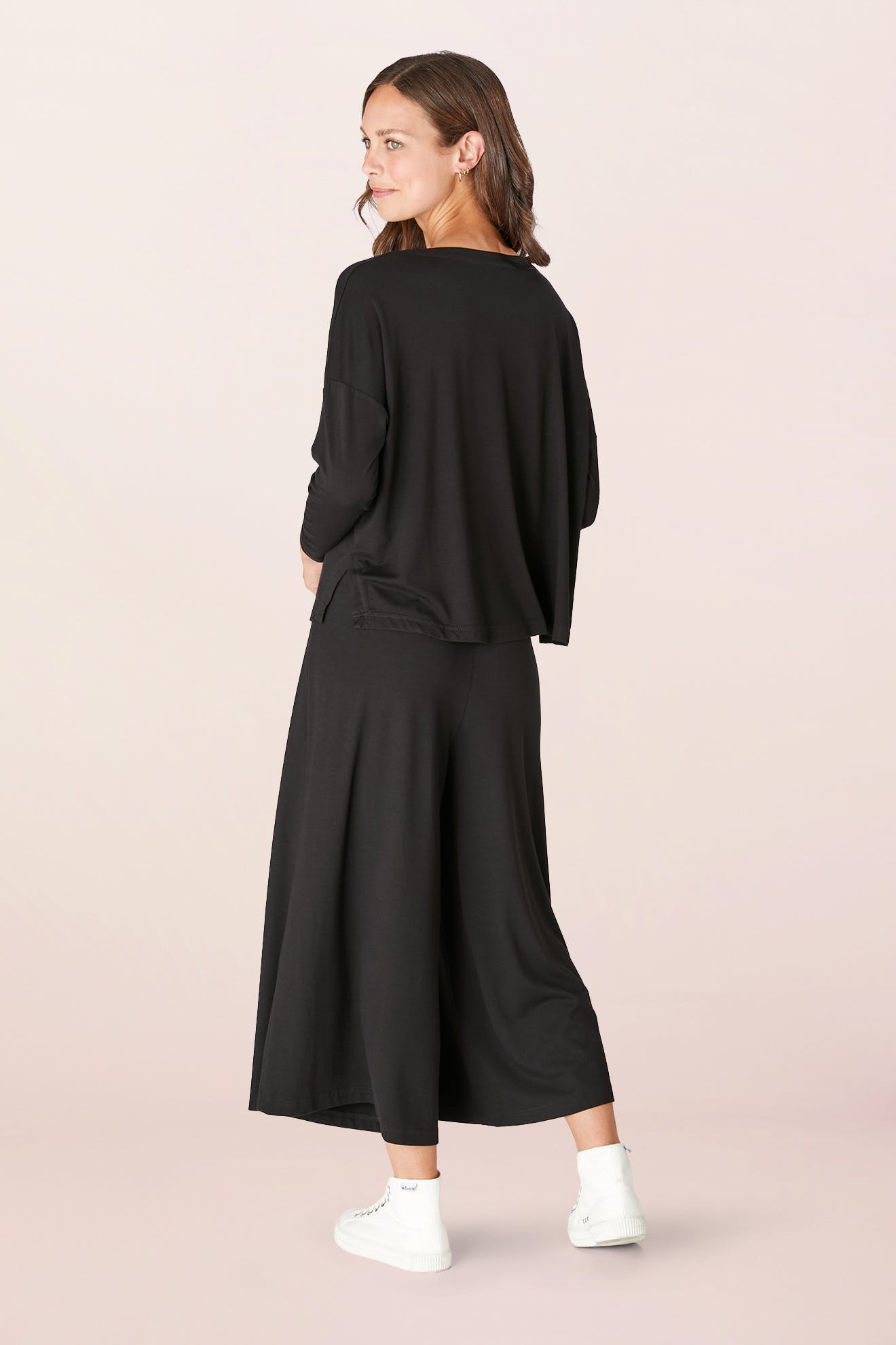 A person wearing a Fluid Crepe Jersey Relaxed Top in black and wide-leg pants, standing in white sneakers against a plain background, embodies an effortless A-line silhouette.