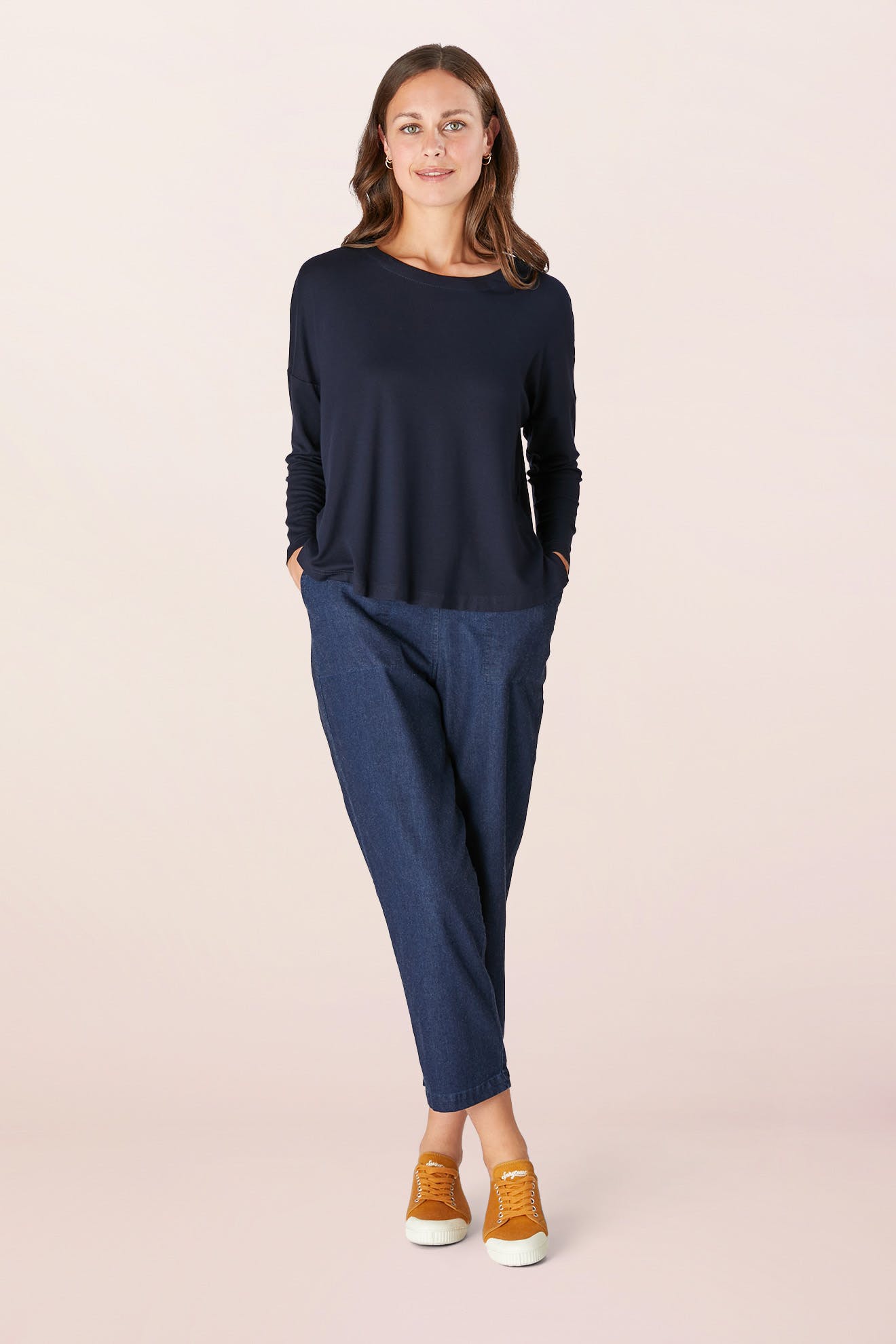 A person stands facing forward with hands in pockets, wearing a relaxed Fluid Crepe Jersey black long-sleeve top, blue pants, and orange sneakers against a plain background.