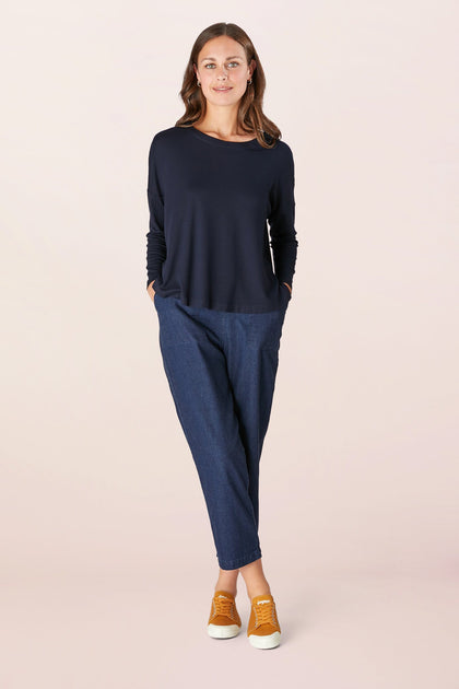 A person stands facing forward with hands in pockets, wearing a relaxed Fluid Crepe Jersey black long-sleeve top, blue pants, and orange sneakers against a plain background.