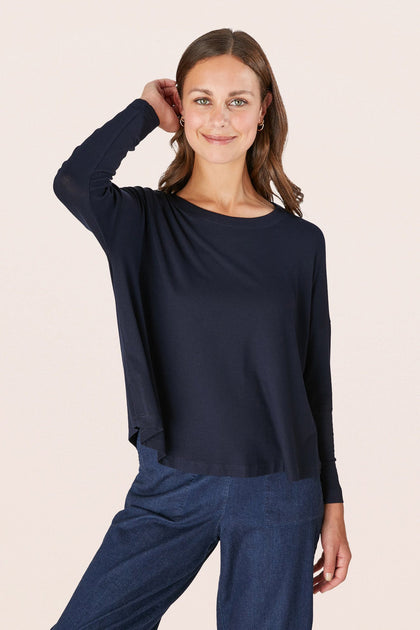 A person is wearing a Fluid Crepe Jersey Relaxed Top in navy paired with blue pants, smiling with one hand on their head against a plain background.