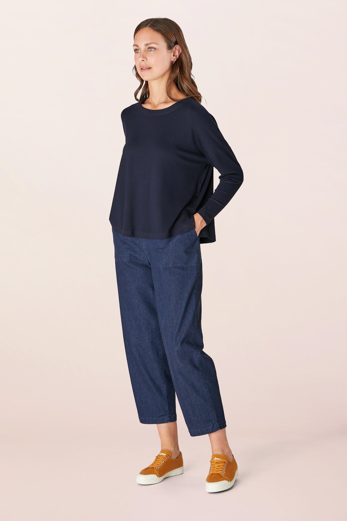 A person wearing a Fluid Crepe Jersey Relaxed Top in navy, blue jeans, and yellow sneakers stands against a plain background.
