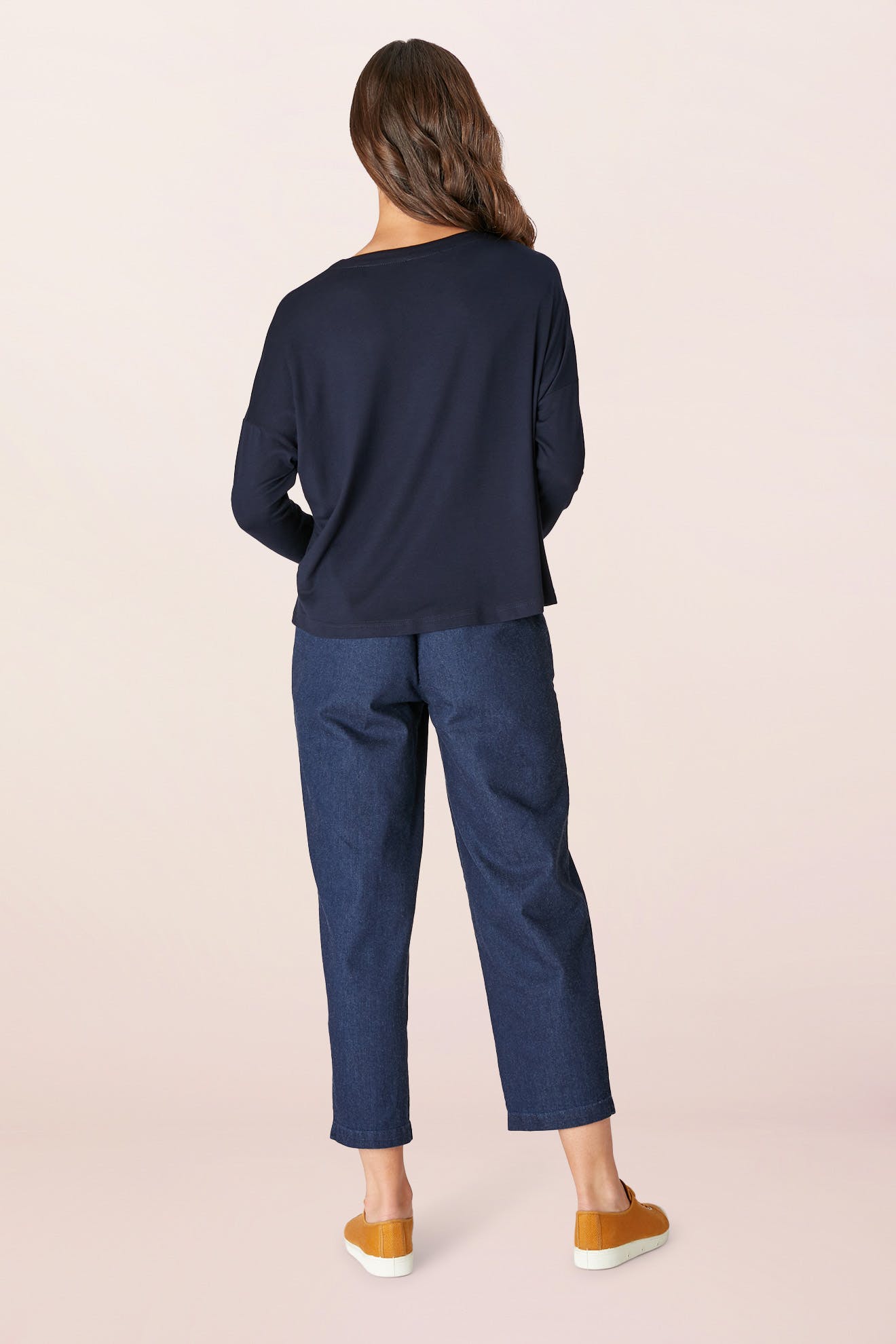 A person with long hair stands facing away, wearing a Fluid Crepe Jersey Relaxed Top in navy, paired with blue jeans and tan shoes, set against a plain background.