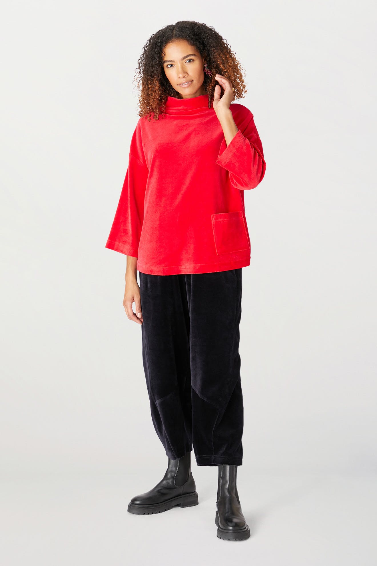 A person stands wearing a Velvet Jersey Cowl Neck Top in red with a pocket, along with black pants and matching black boots, set against a plain background. Ideal for the holiday party season!