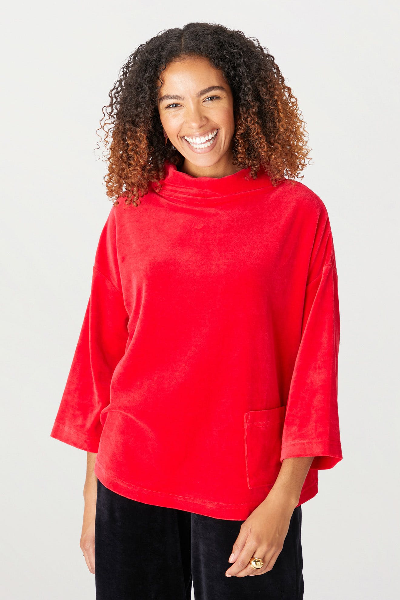 A person with curly hair, wearing a red velvet jersey cowl neck top with a front pocket and a swingy silhouette, smiles while standing against a plain background.