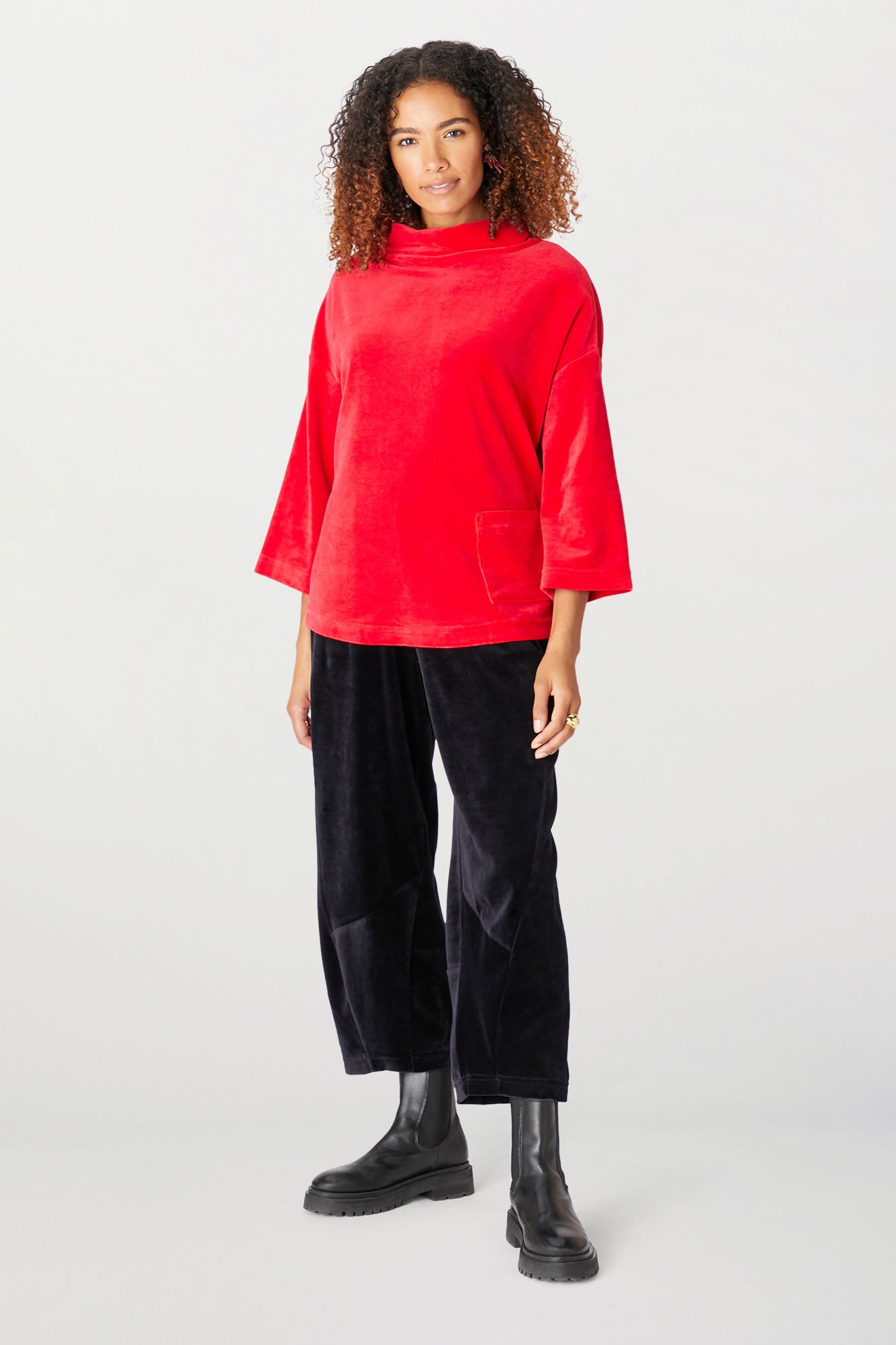 A person stands against a plain background wearing a red Velvet Jersey Cowl Neck Top, black pants, and black boots. They have curly hair and a neutral expression, perfectly capturing the essence of the holiday party season.