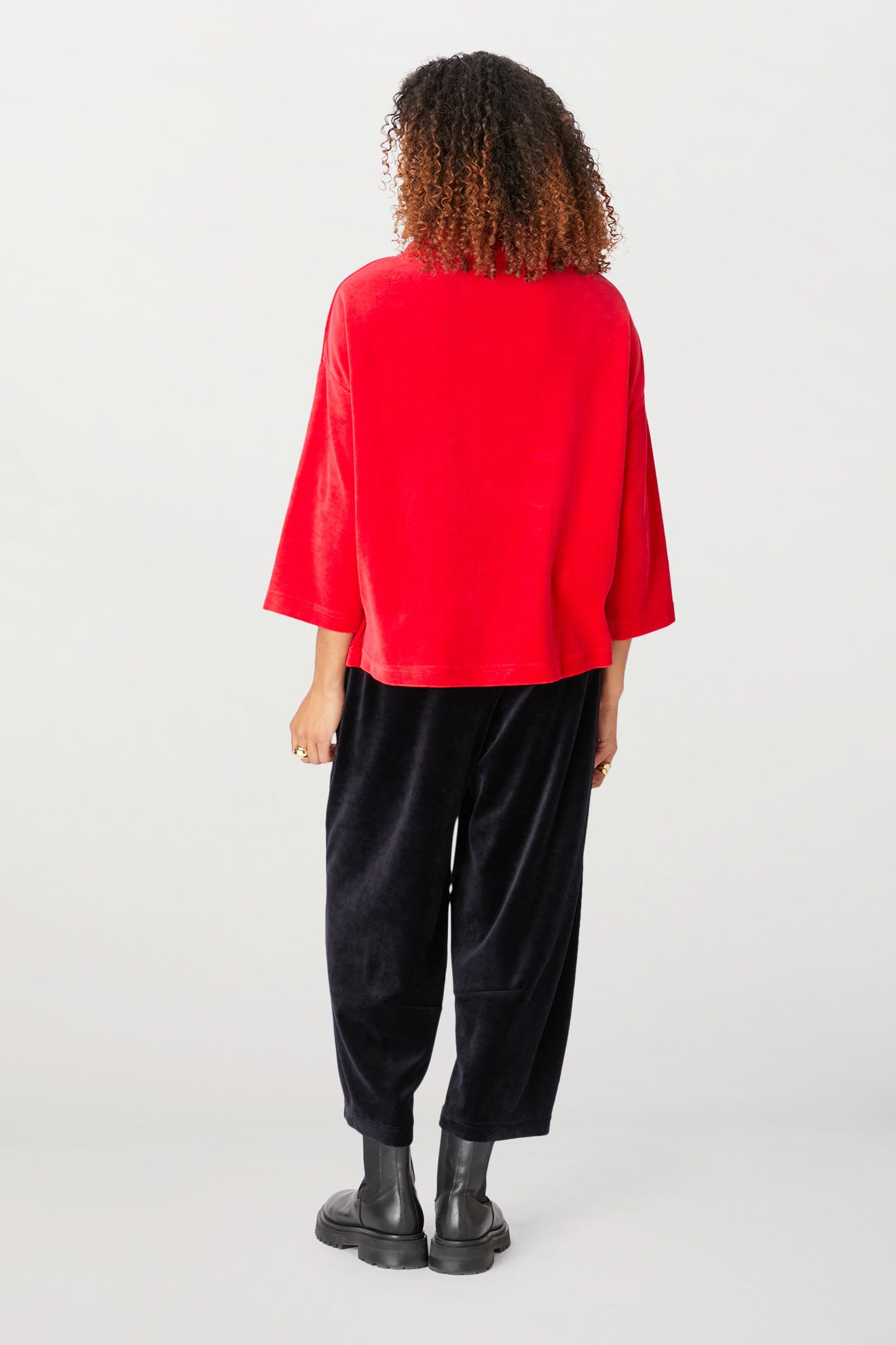 A person with curly hair, wearing a red Velvet Jersey Cowl Neck Top, black loose-fitting pants, and black boots, stands facing away from the camera.
