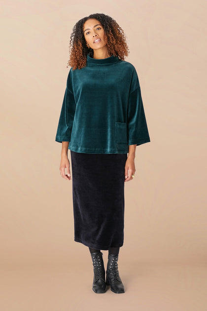 A woman with curly hair is wearing a dark green Velvet Jersey Cowl Neck Top, a black skirt with a swingy silhouette, and black ankle boots. She stands against a beige background. This outfit is perfect for the holiday party season.