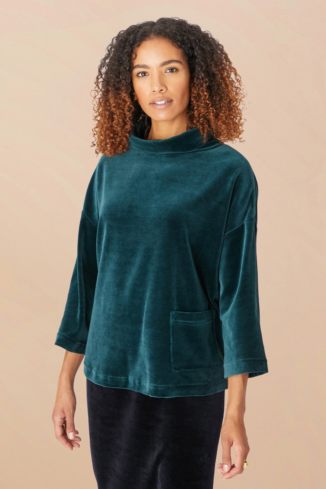 A woman with curly hair stands against a beige background, donning a teal Velvet Jersey Cowl Neck Top with three-quarter sleeves, perfectly poised for the holiday party season.