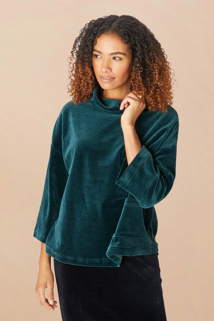 A woman stands against a plain background, wearing a dark green Velvet Jersey Cowl Neck Top with long sleeves. She has curly hair and is looking to the side. The swingy silhouette of her attire perfectly captures the festive spirit of the holiday party season.