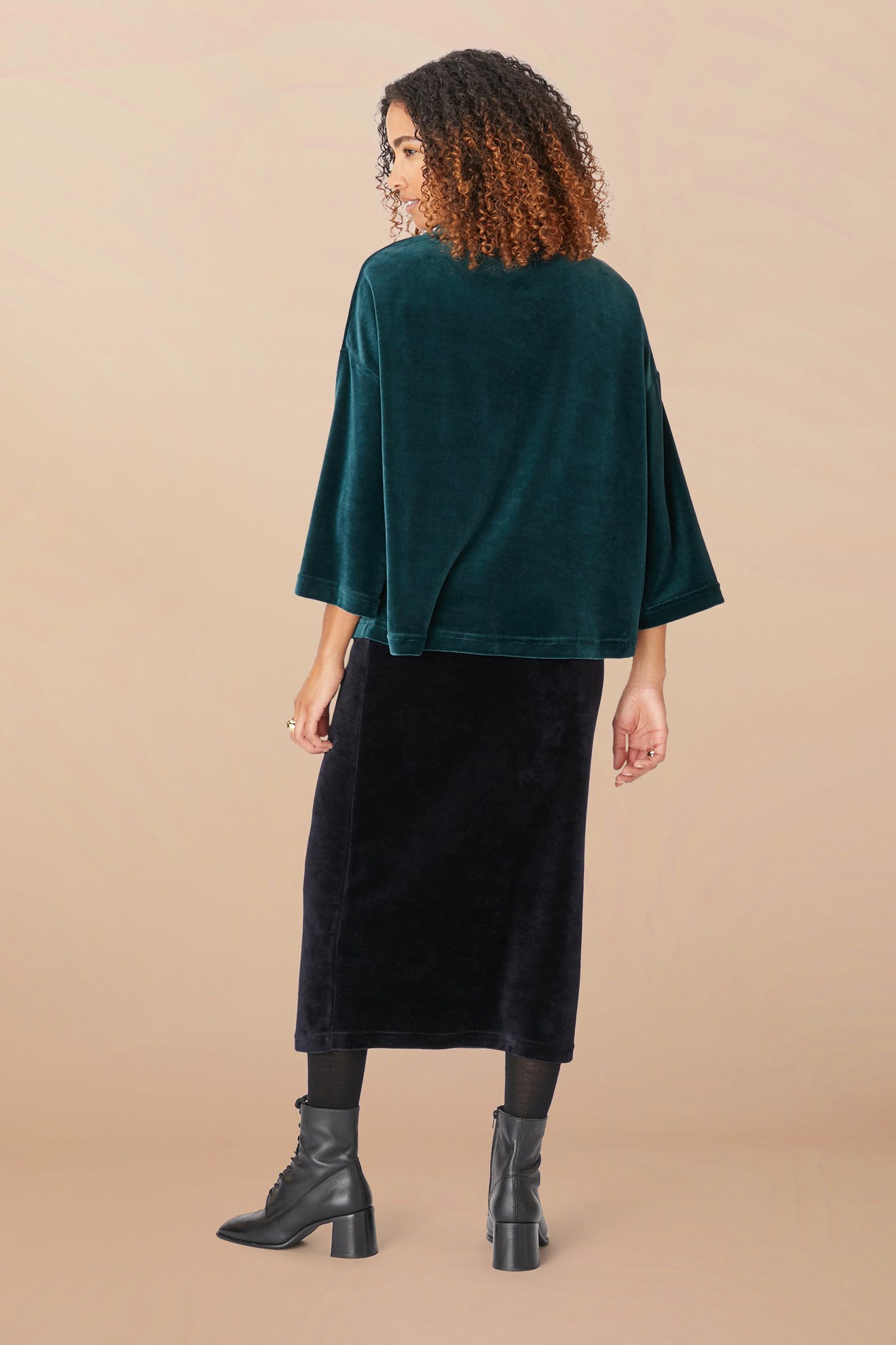 A woman with curly hair wearing a green Velvet Jersey Cowl Neck Top, a black skirt, and black boots is standing turned away from the camera against a plain beige background. The swingy silhouette of her outfit perfectly captures the essence of holiday party season.