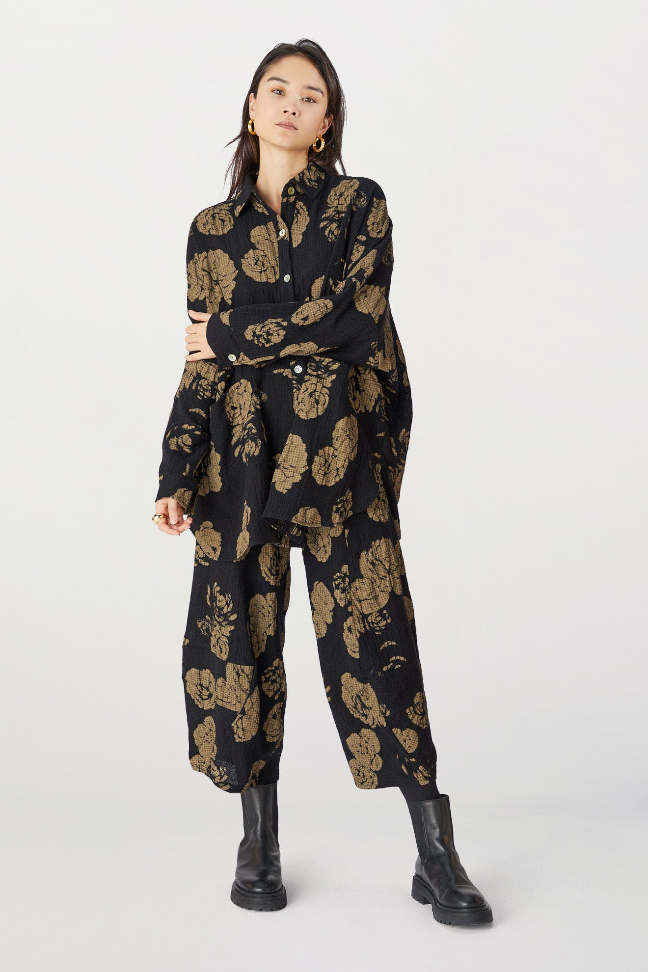A woman in dark Rose Print Pants with black boots stands with arms crossed against a light background.