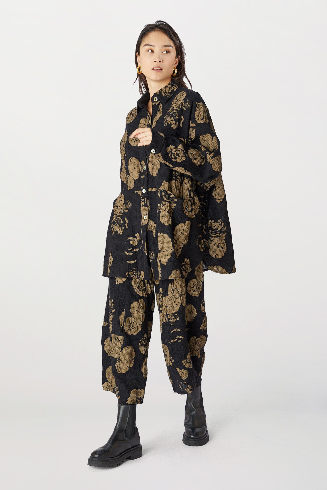 A woman wearing a black Rose Print Pant jacket and matching pants with black boots, standing against a light background.