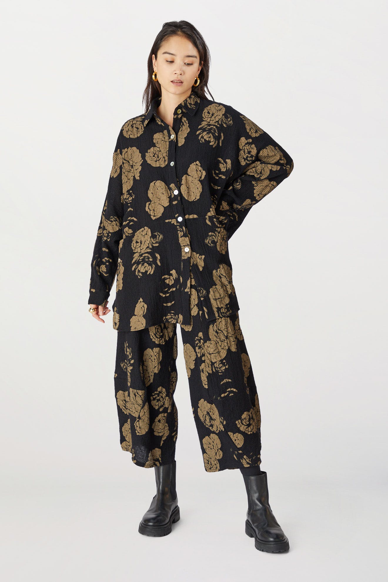 A young woman stands wearing a black and gold rose print pant with an elasticated waist and button-up front, paired with black boots, against a light background.