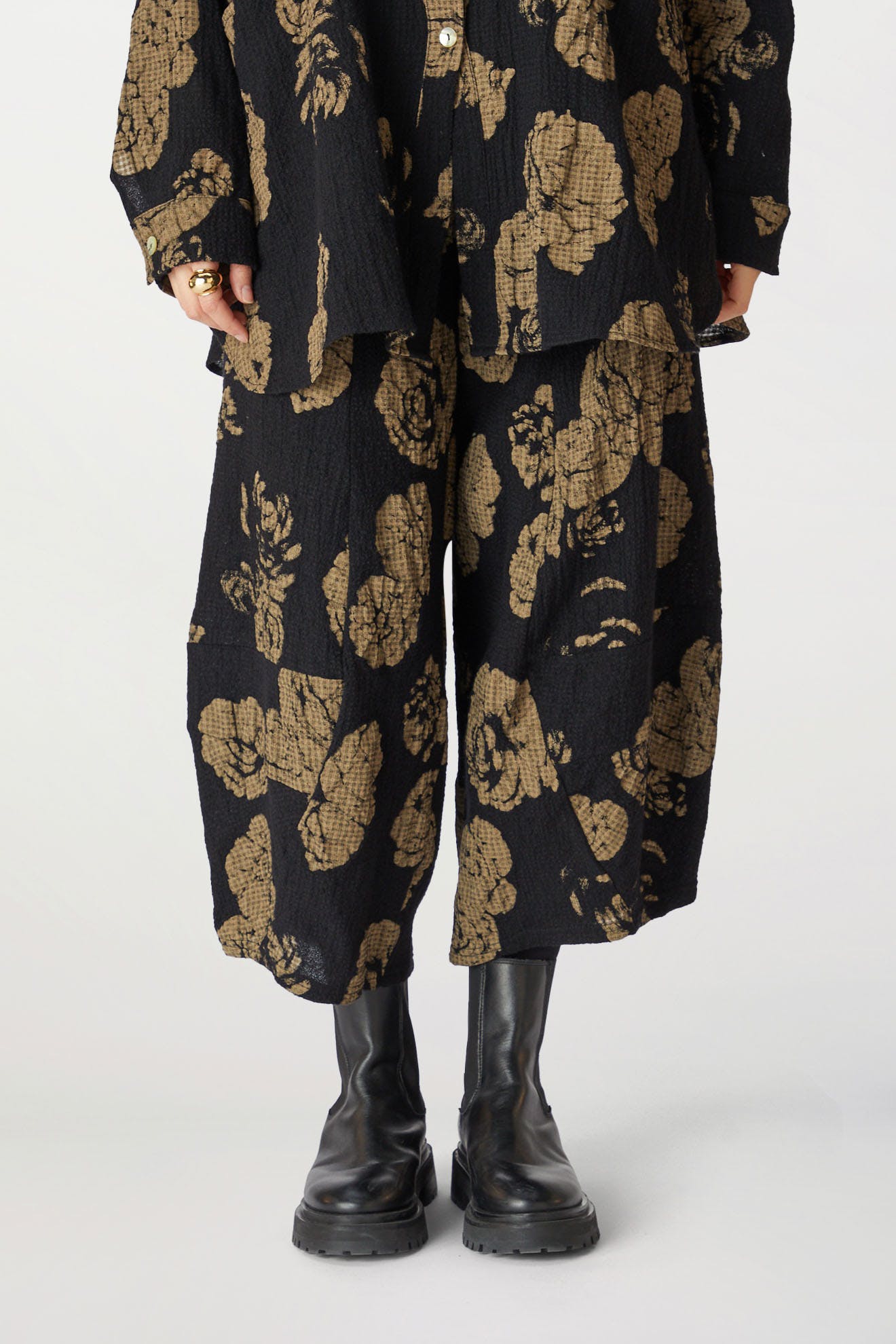 Person standing in Rose Print Pant and gold pants and matching jacket, hands partially concealed, wearing black boots.