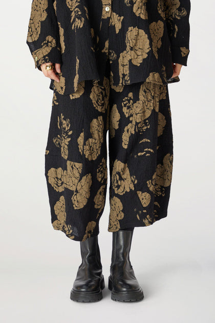 Person standing in Rose Print Pant and gold pants and matching jacket, hands partially concealed, wearing black boots.
