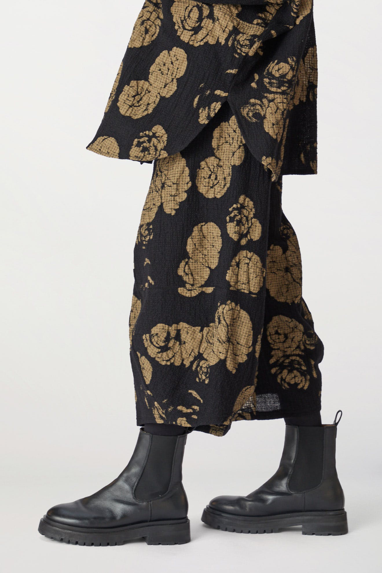 Close-up view of a person wearing black boots and a draped rose print pant with an elasticated waist in black and gold tones.
