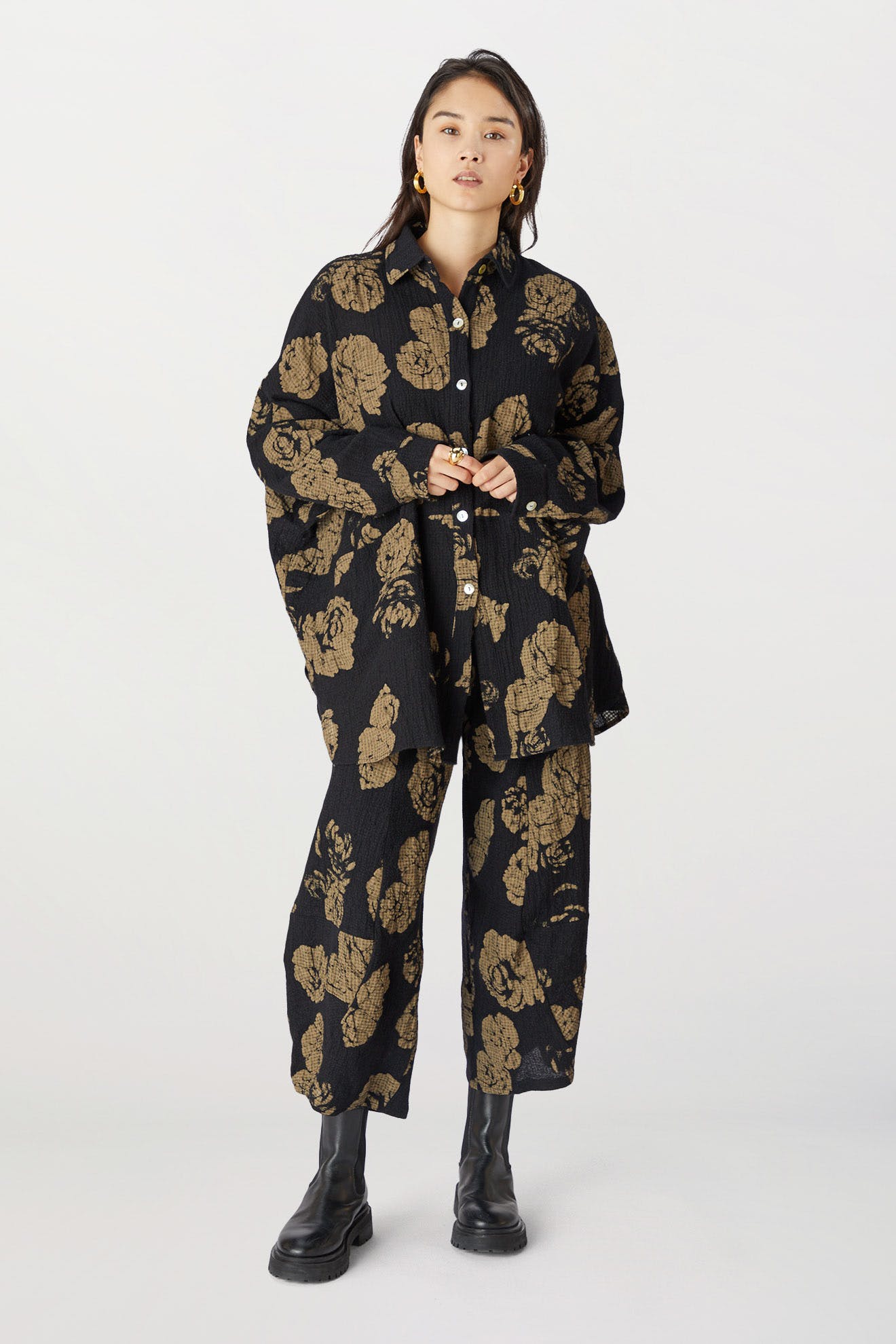 Woman standing in a studio, wearing a black oversized shirt and Thanny trousers with a golden rose print pant, paired with black boots.