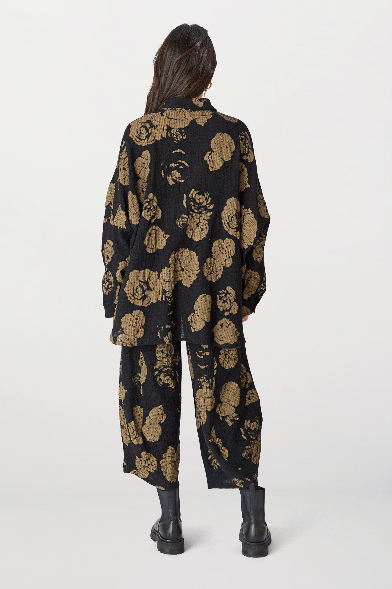 A woman from behind wearing black oversized Rose Print Pant, paired with black boots, standing against a white background.