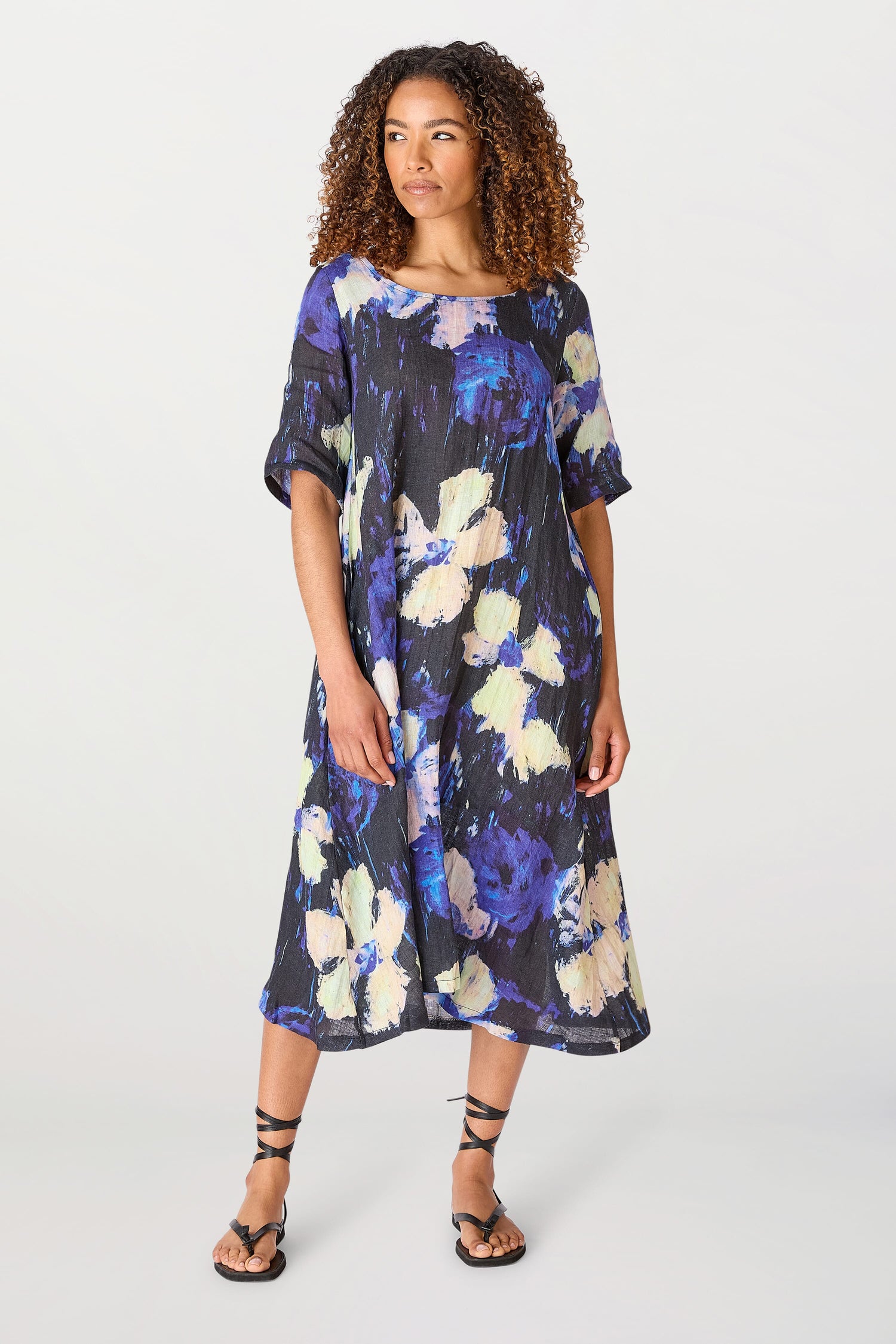 A woman with curly hair stands in a studio, wearing a Floral Linen Dress and black sandals.