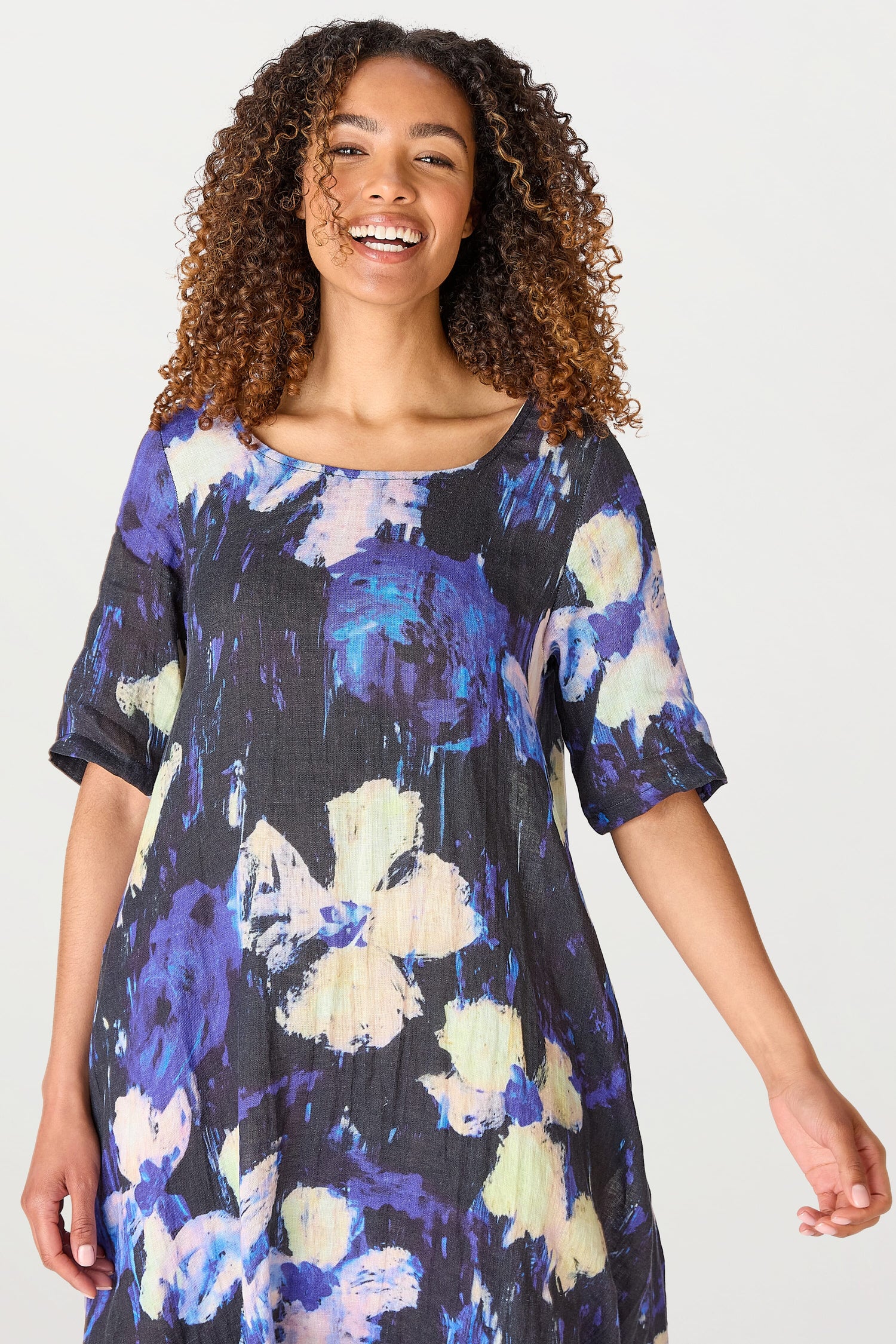 A smiling woman with curly hair, wearing a Floral Linen Dress, standing against a light background.