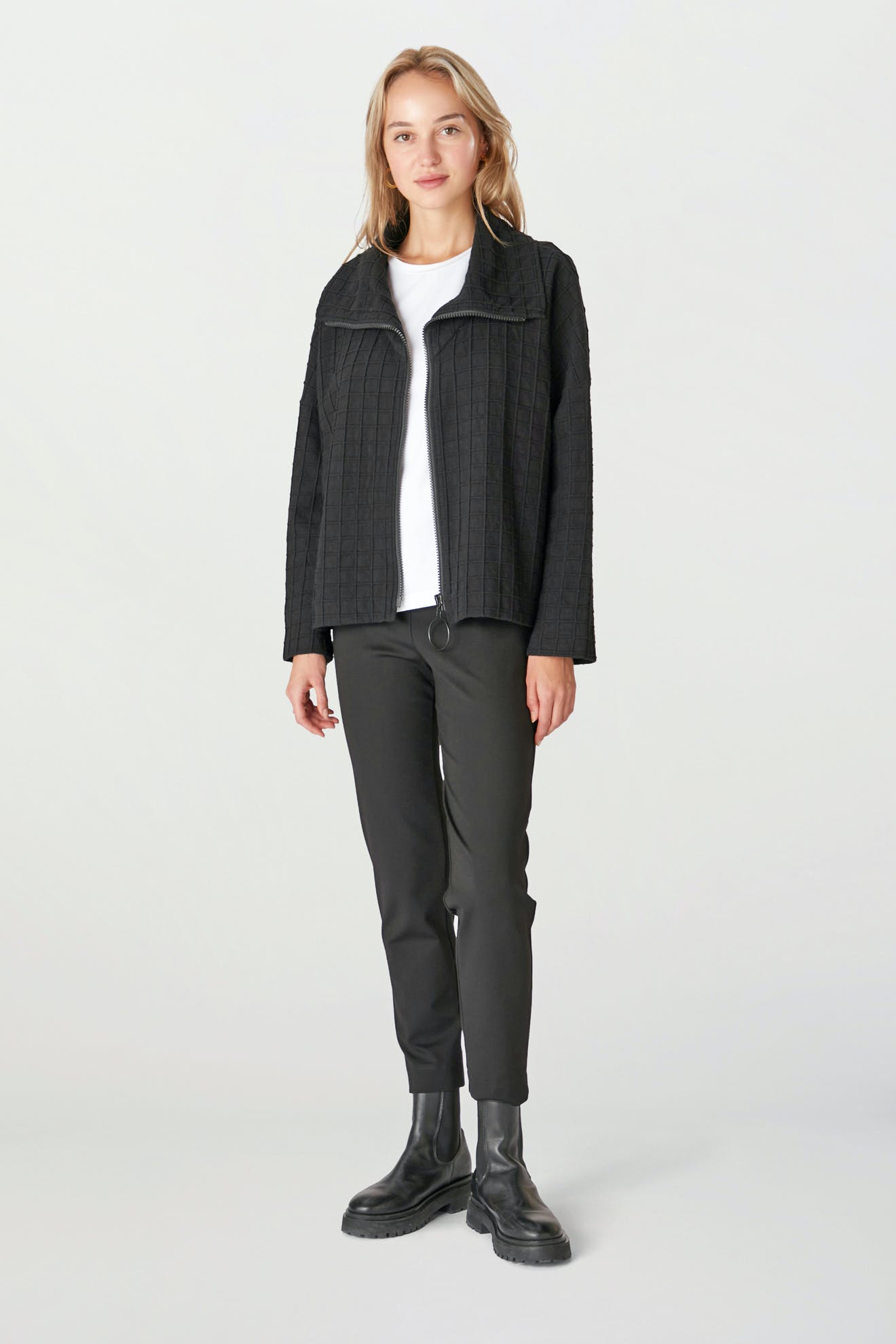 A person stands wearing the Window Pane Wool Jacket, featuring a structured black design and an oversized folded collar, complemented by a white shirt, black pants, and black boots against a plain background.