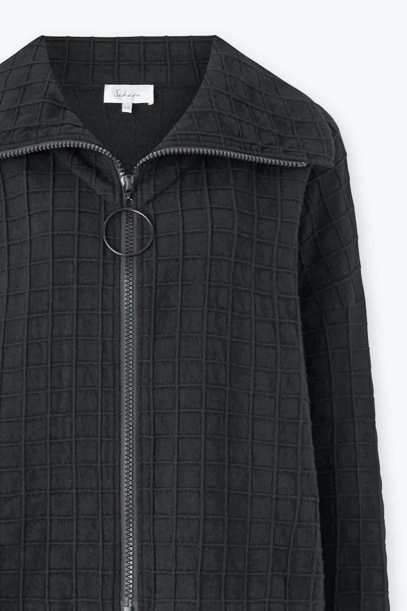 Close-up of a Window Pane Wool Jacket, a black cotton blend with a grid pattern. It showcases an oversized folded collar, a circular zipper pull, and a visible clothing label inside the collar.