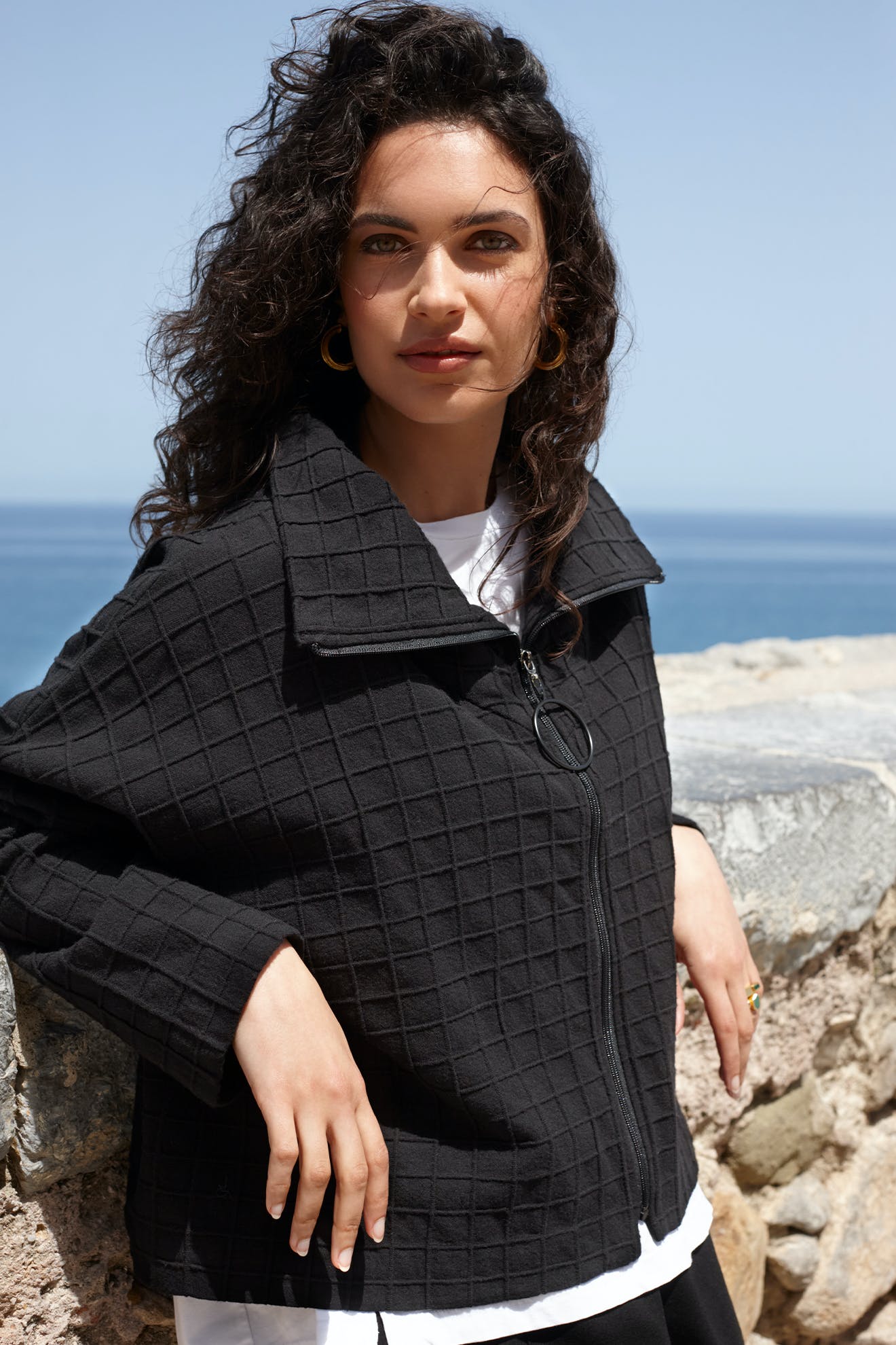 A person with curly hair stands outdoors against a backdrop of the ocean and sky, wearing the chic Window Pane Wool Jacket. Its oversized folded collar adds a stylish flair to the scenic view.